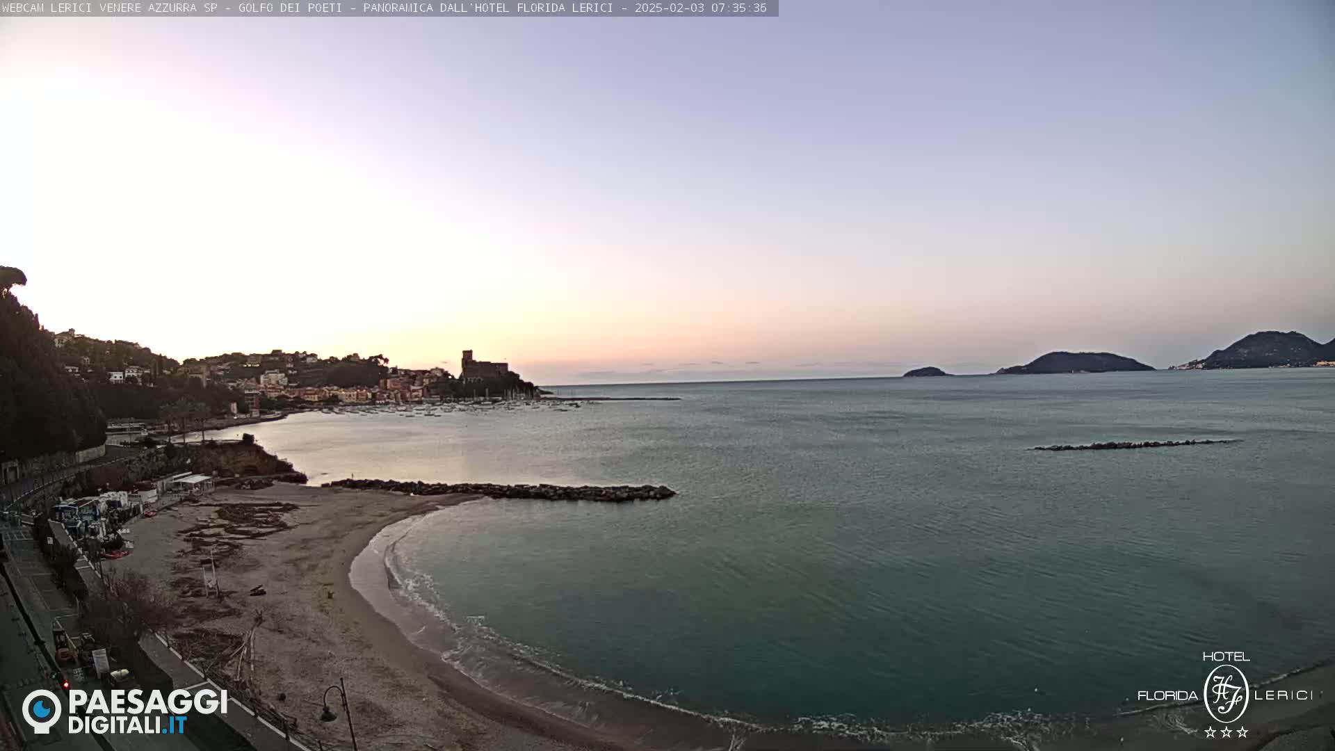 A tranquil coastal scene at dawn shows a sandy beach curving into a calm sea, with a town nestled against hills in the background, under a pale, clear sky.