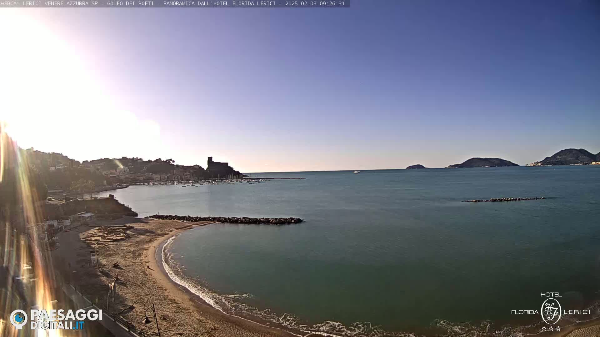 A sandy beach curves into a calm, teal-colored sea, with a town and castle visible in the background under a clear, bright sky.