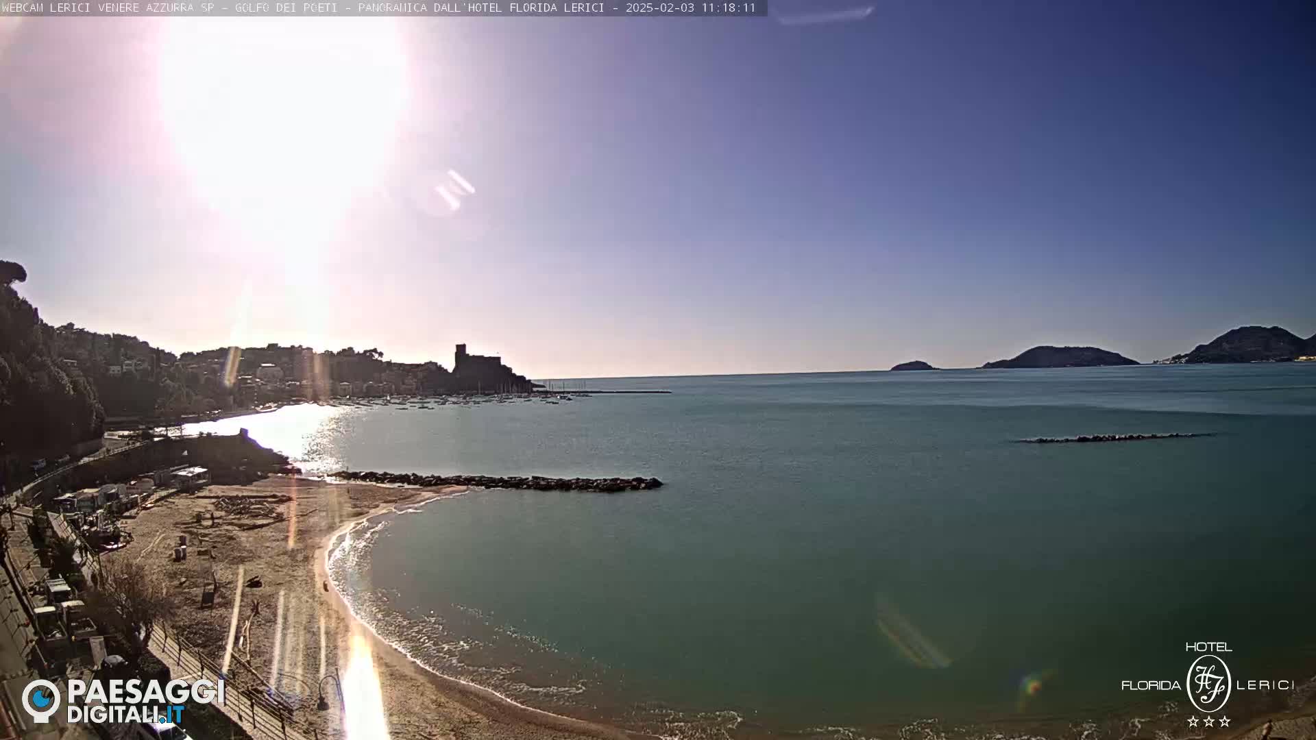 A sandy beach curves into a calm, teal sea, with a town and a distant hill visible under a sunny sky.