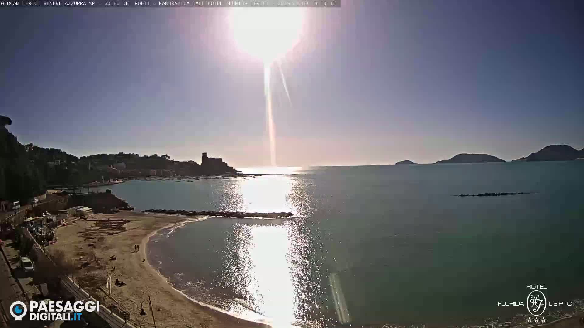 A sunny day at a beach with calm, sparkling water, showing a sandy shore, some buildings, and hills in the distance.