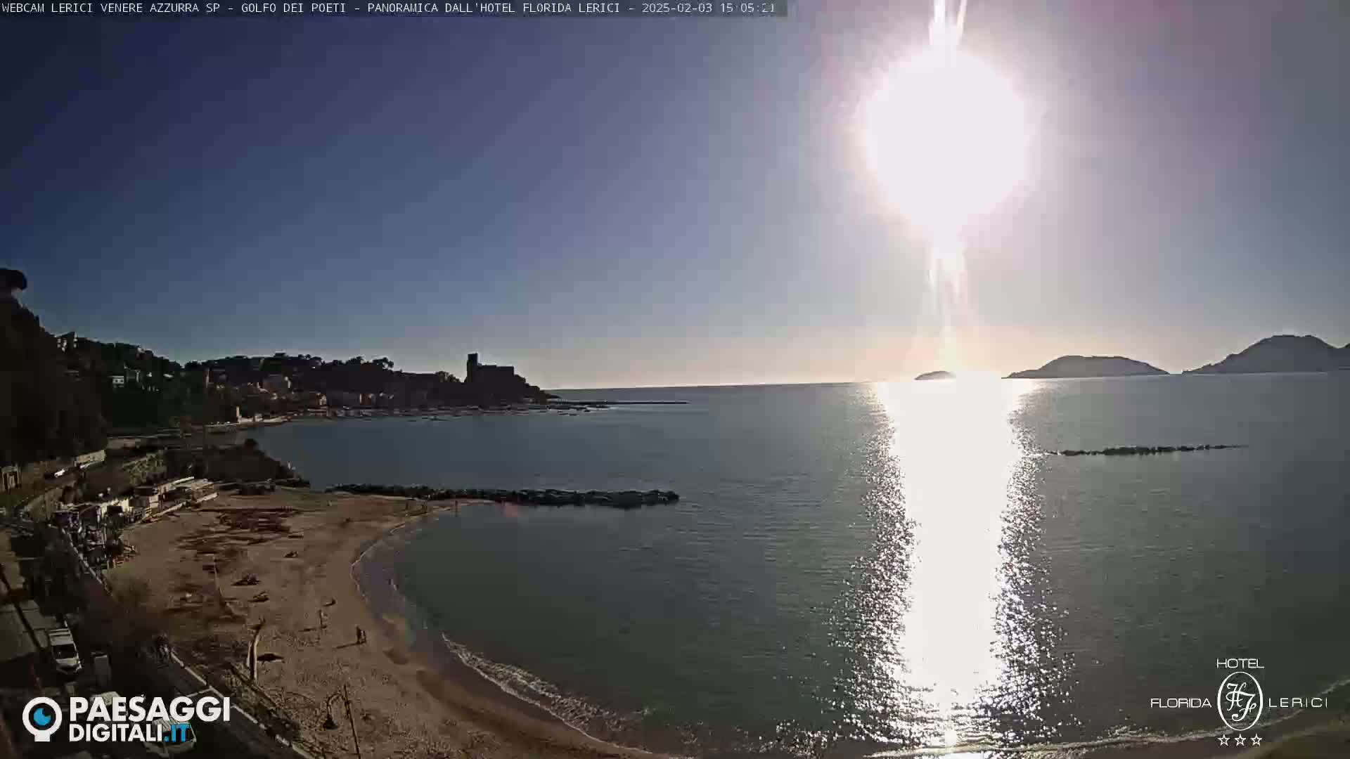 A sunny day shows a sandy beach, calm ocean waters, and distant hills with a town visible in the background.