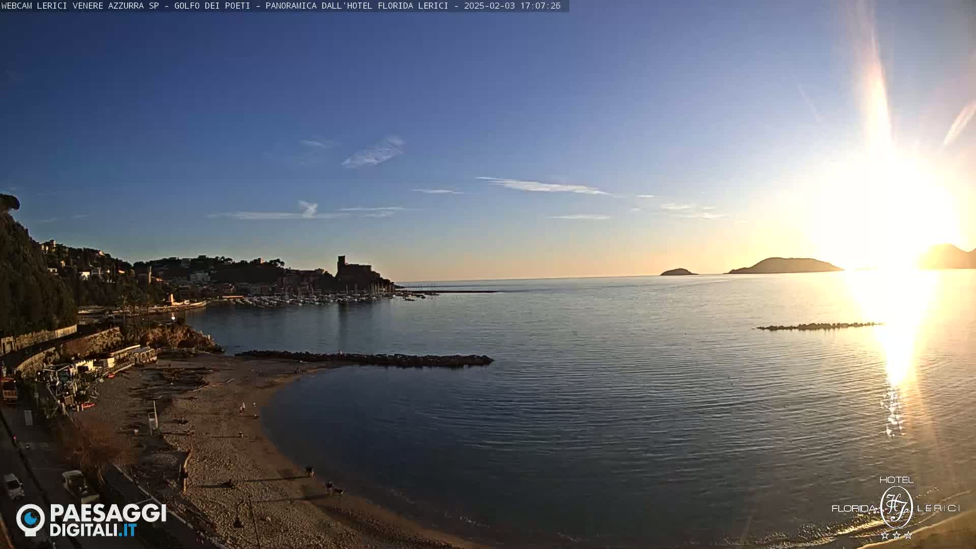A coastal town with a harbor is seen at sunset, with a sandy beach in the foreground and calm water reflecting the bright sun.