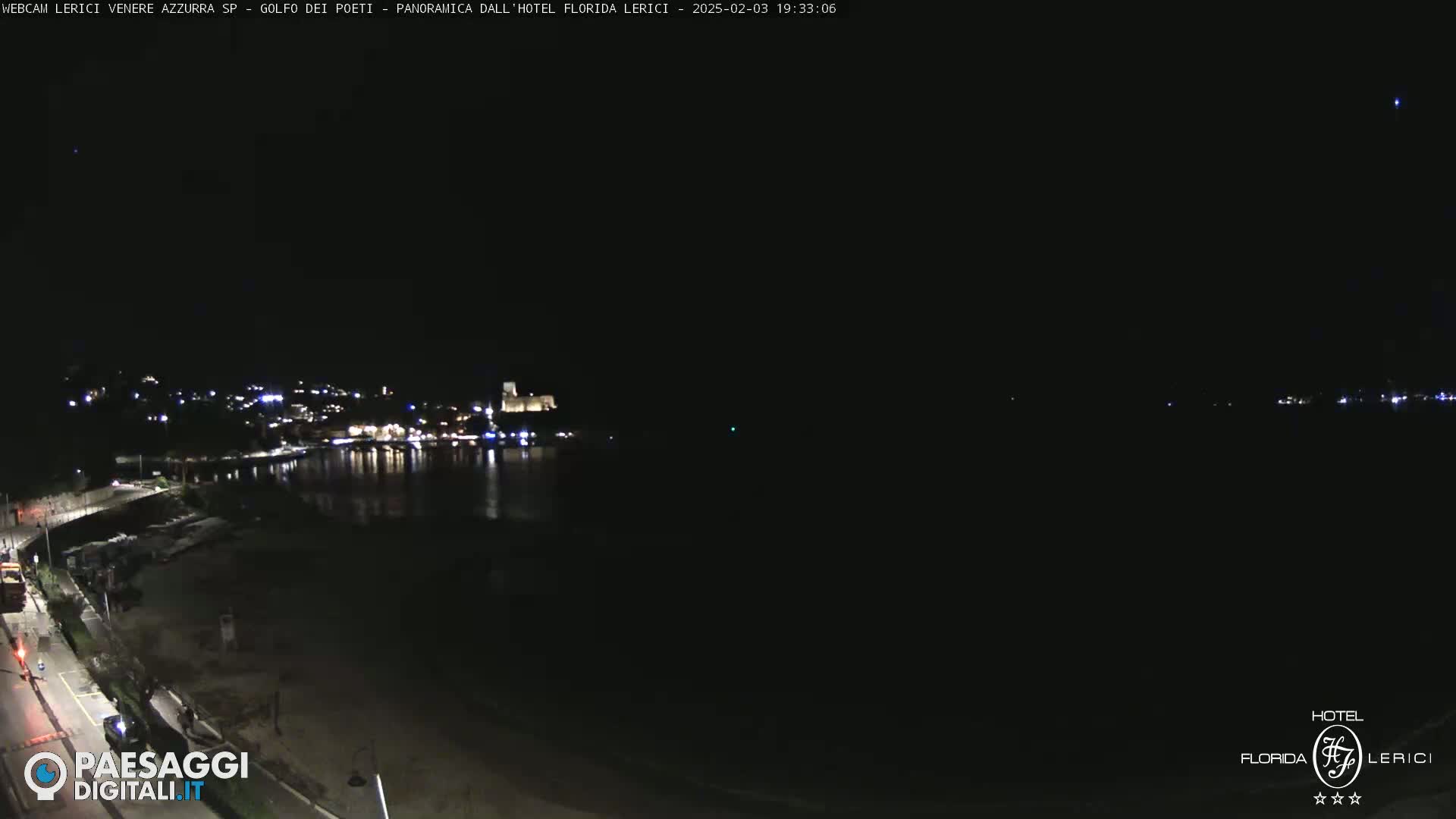 A nighttime view of a dark coastline with a town’s lights visible across the water.