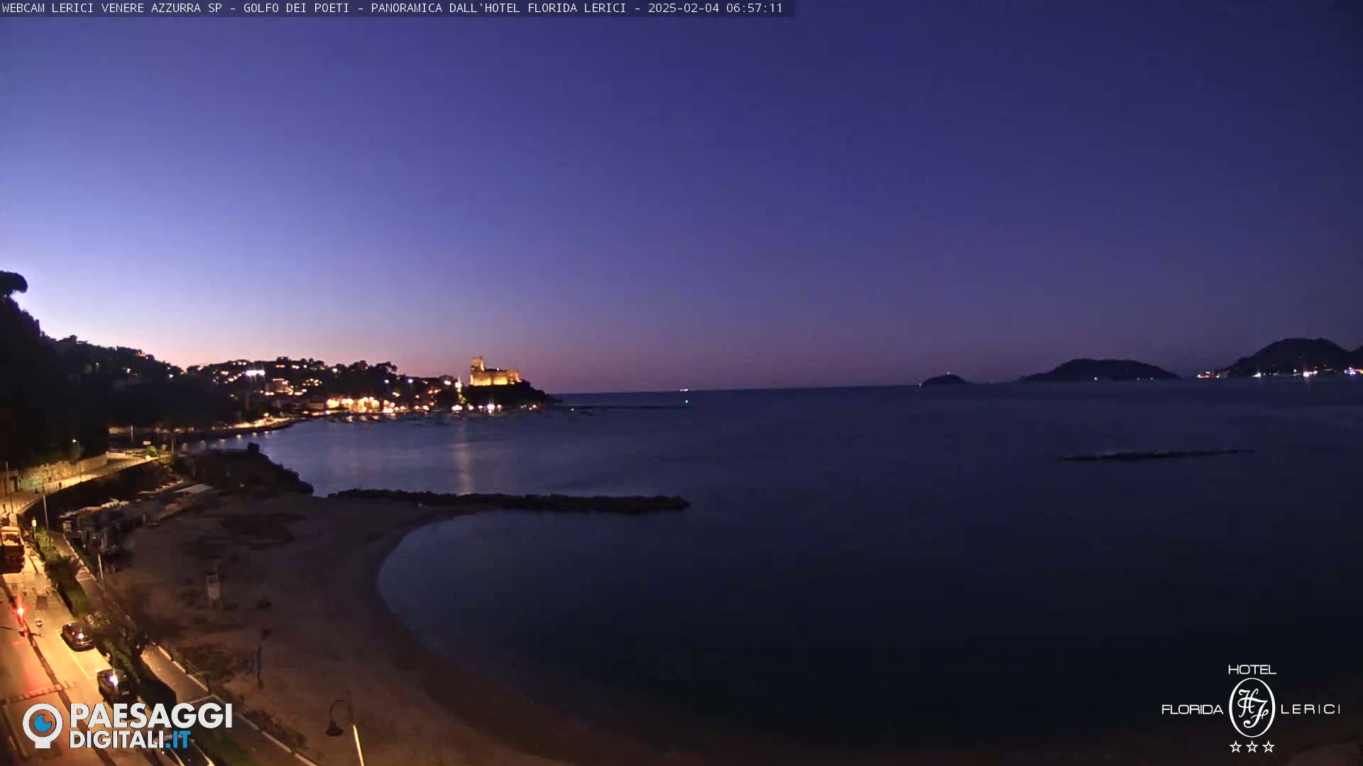 A nighttime coastal scene shows a calm, dark-blue sea under a twilight sky, with a town and castle illuminated on the left shore and darker hills in the distance.