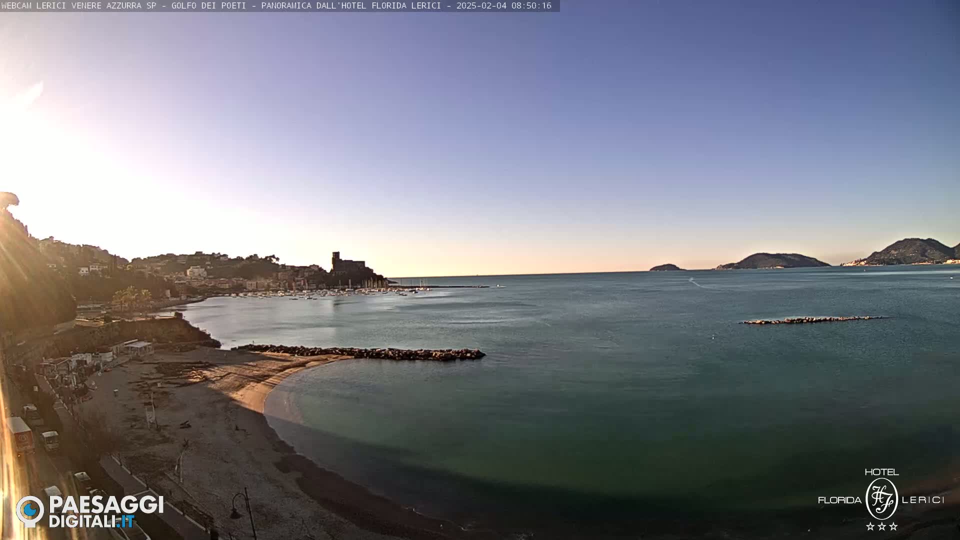 A clear, sunny morning shows a calm sea with a sandy beach, a town nestled against a hillside, and distant islands under a pale blue sky.