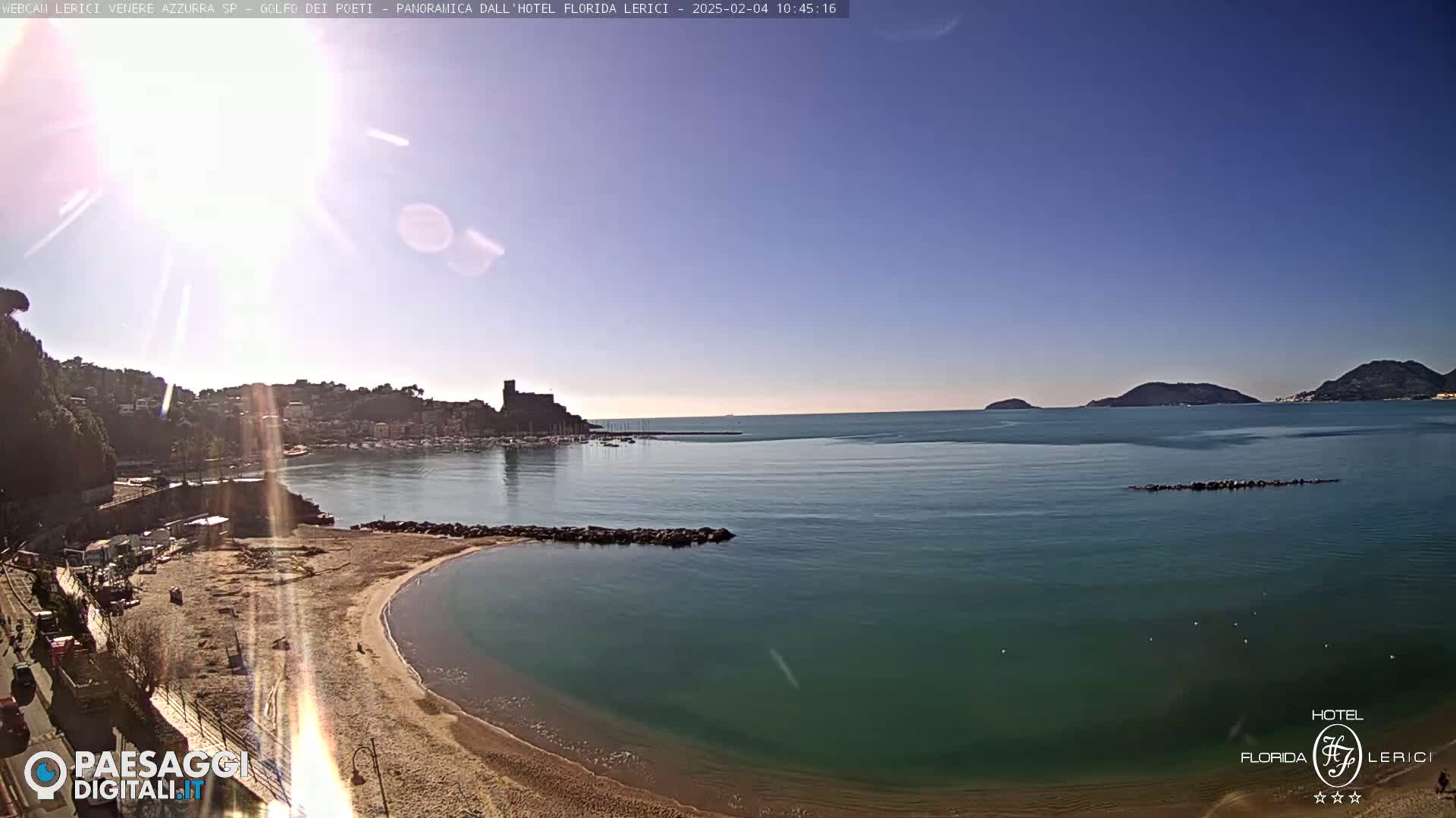 A sandy beach curves into a calm, teal sea under a bright, sunny sky, with a town and distant hills visible in the background.