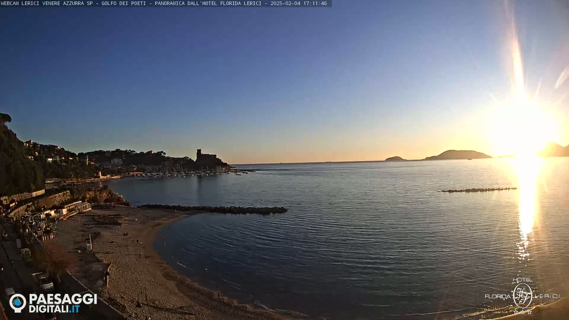 A coastal town with a sandy beach and calm bay is bathed in the golden light of a setting sun under a clear blue sky.