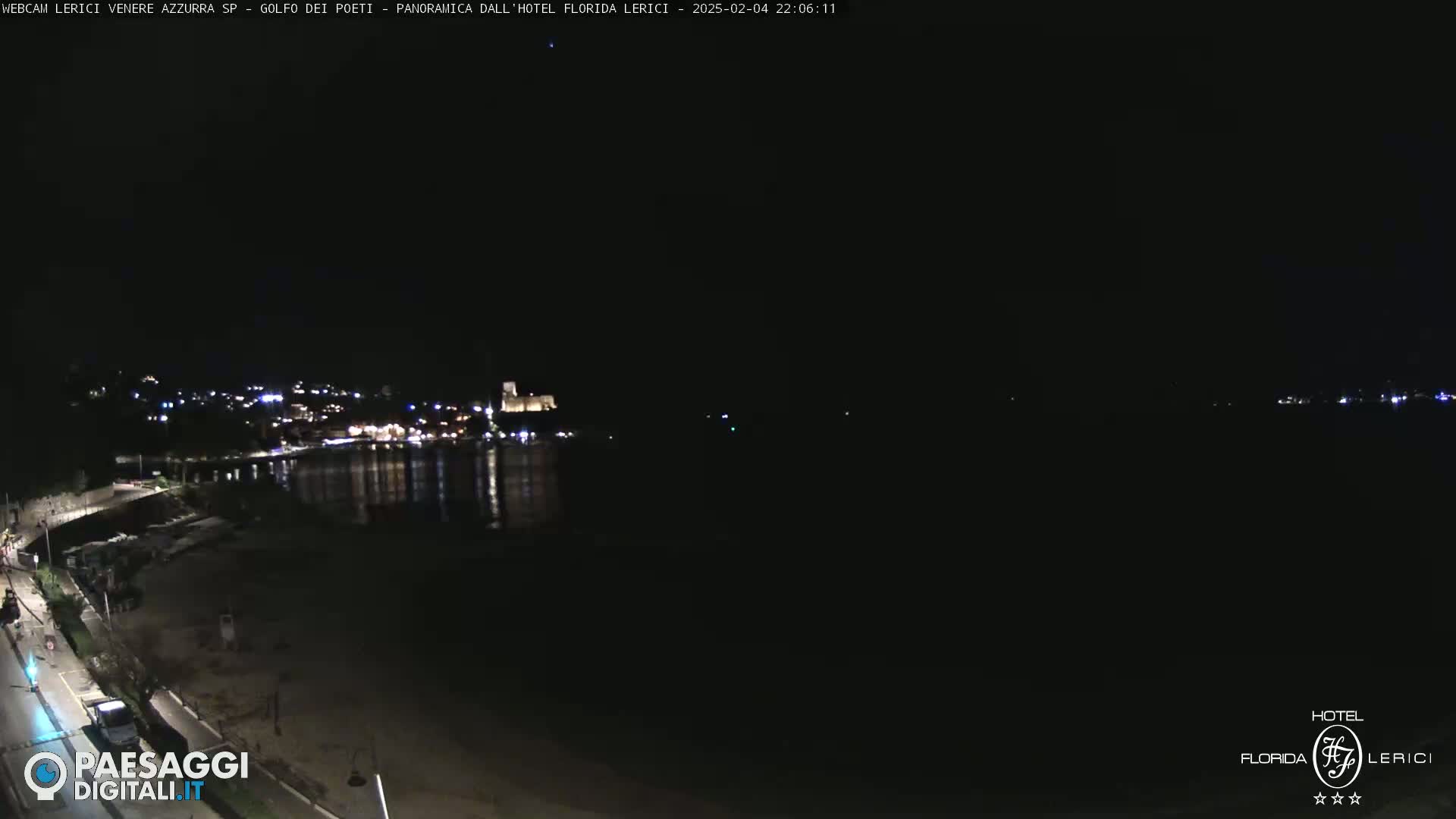A nighttime view of a dark beach and calm water, with a dimly lit town visible across the water.