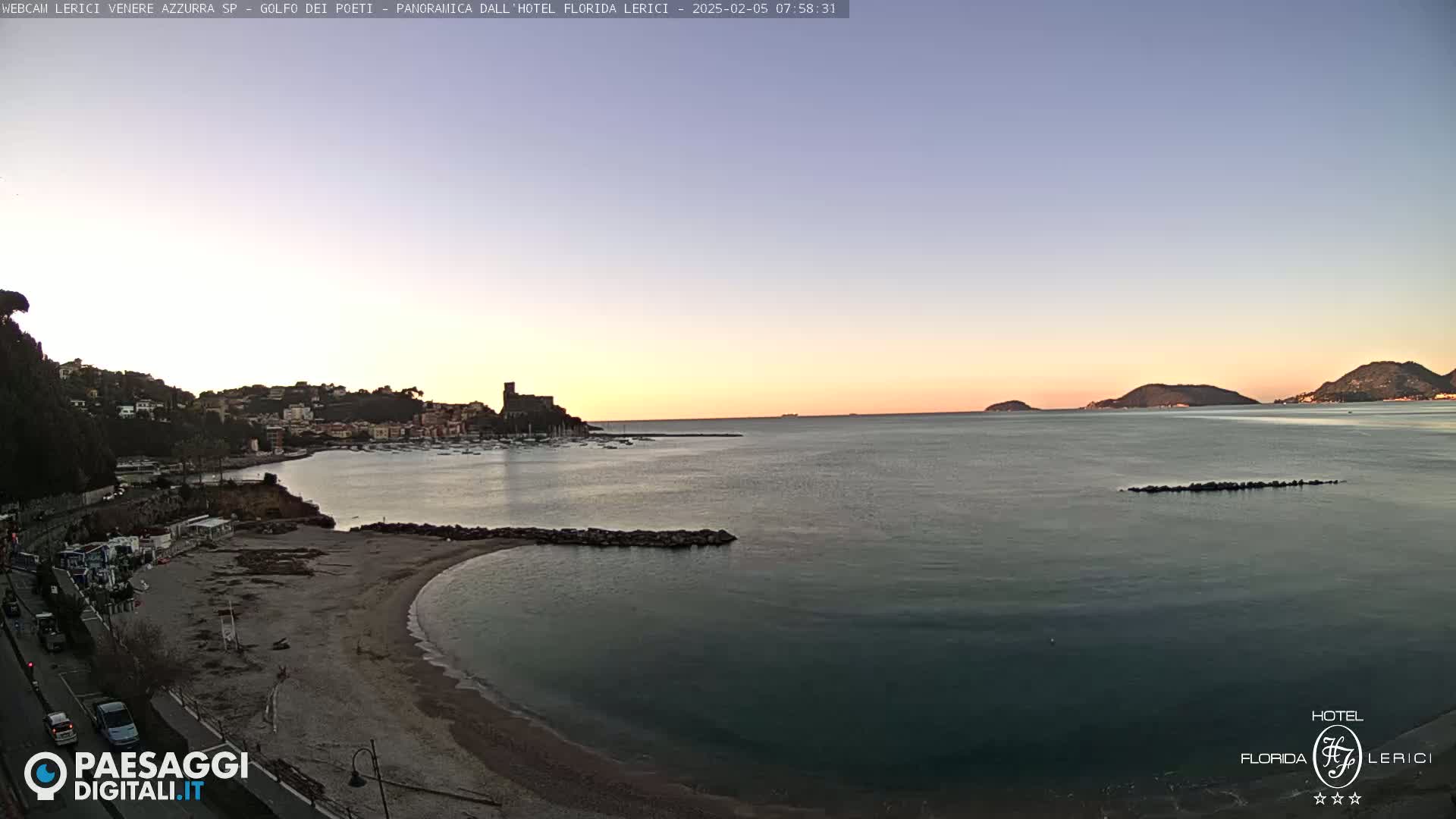 A tranquil seaside town at dawn, under a clear, pastel sky, features a calm bay with a sandy beach and distant hills.