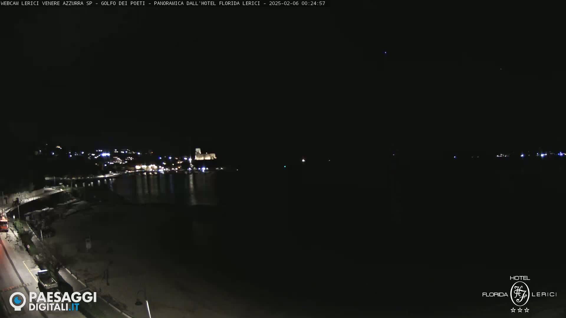 A nighttime long shot shows a dark coastline with a few lights from a town and harbor across a dark body of water.