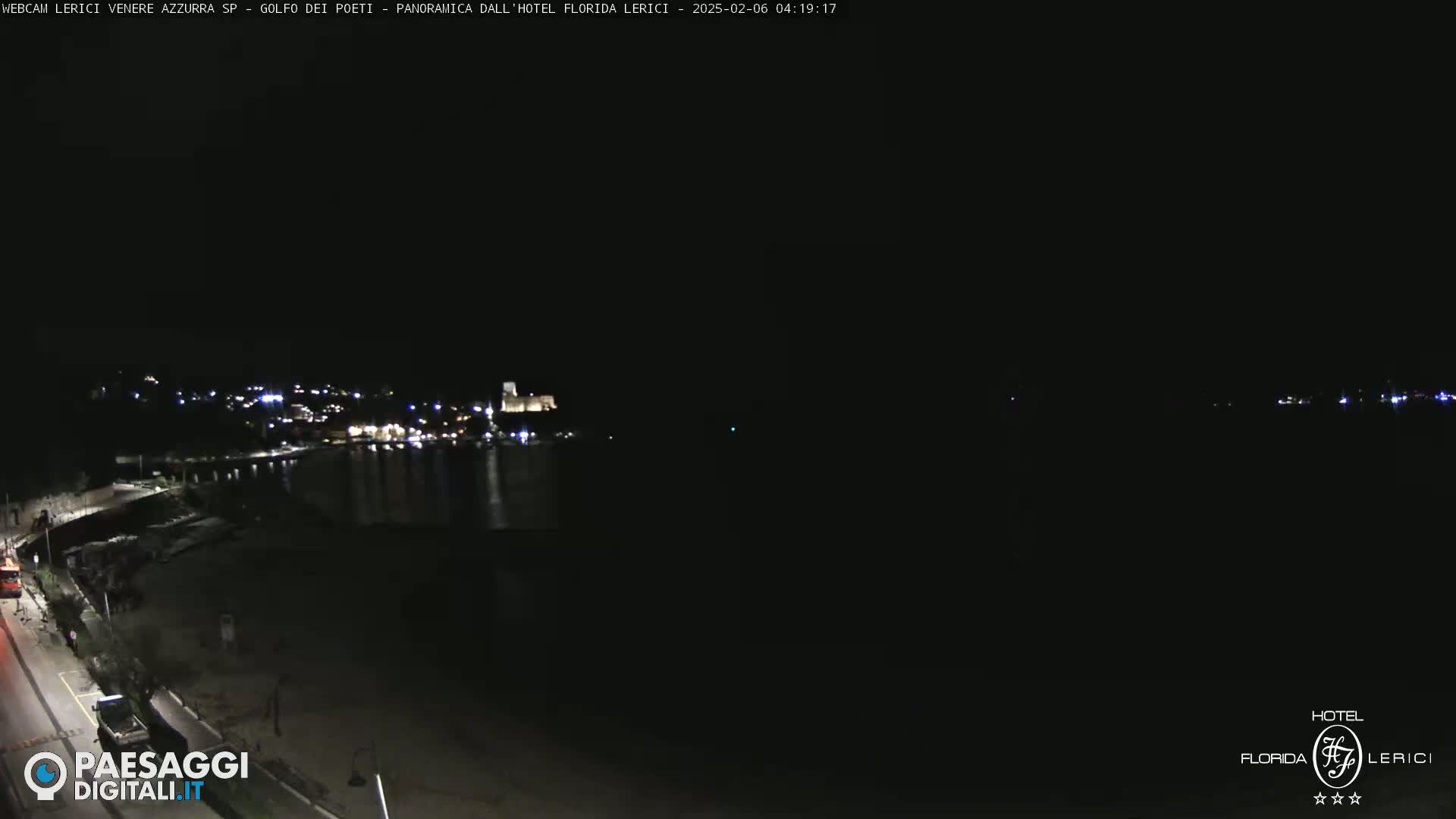 A nighttime, long-range view of a coastal town with lights scattered across the shoreline and a dark body of water.