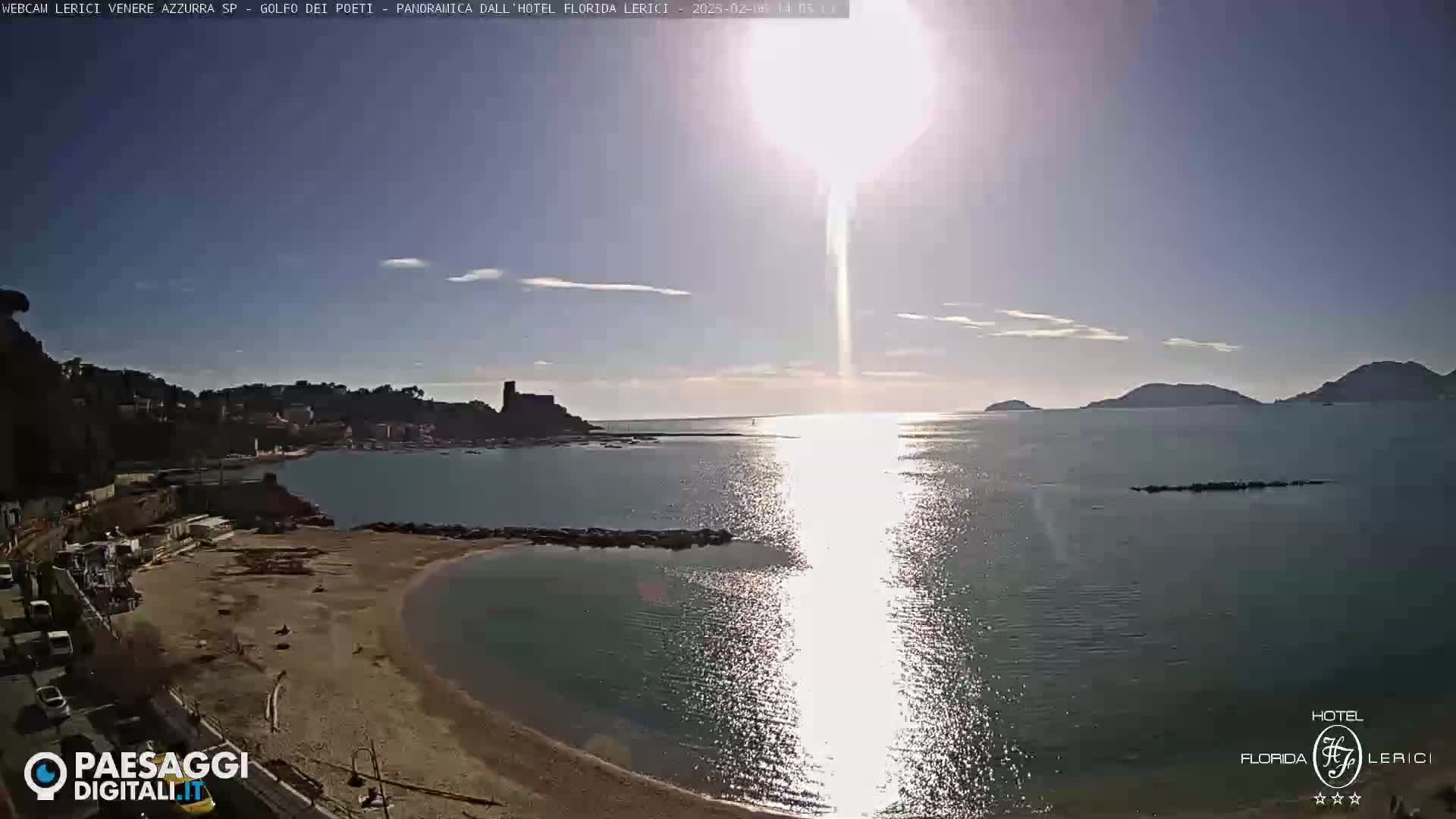 A sandy beach curves into a calm bay, with a town and castle visible across the water under a bright, sunny sky.