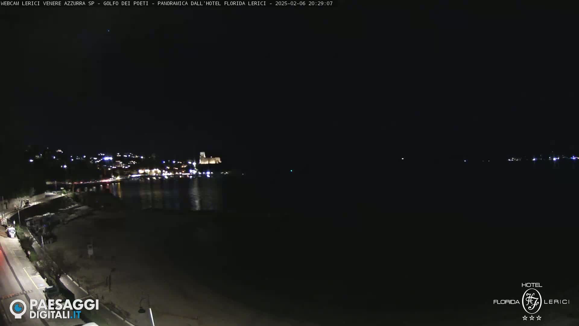 A nighttime coastal scene shows a dark beach and calm water reflecting the lights of a town and a castle in the distance under a clear, dark sky.