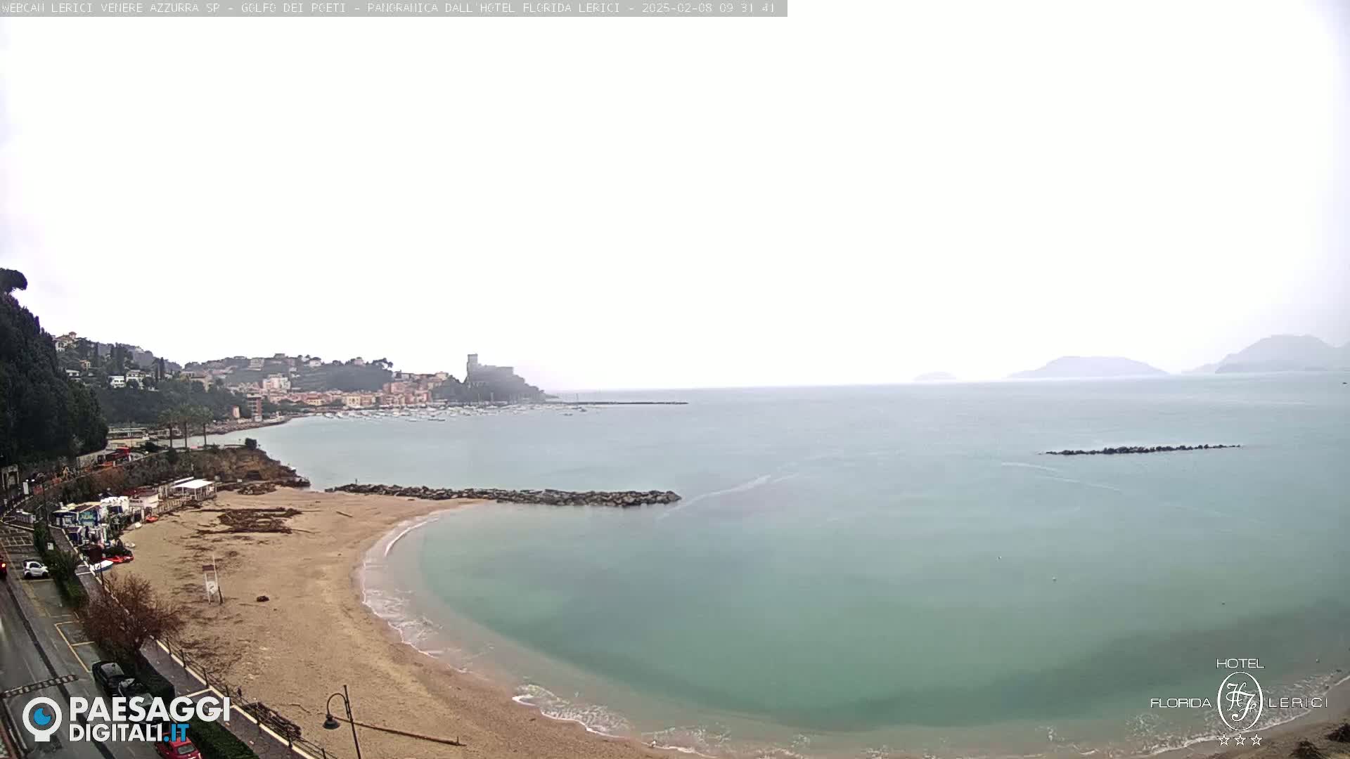 A sandy beach curves into a calm, pale-green sea under an overcast sky, with a town nestled on a hillside in the background.