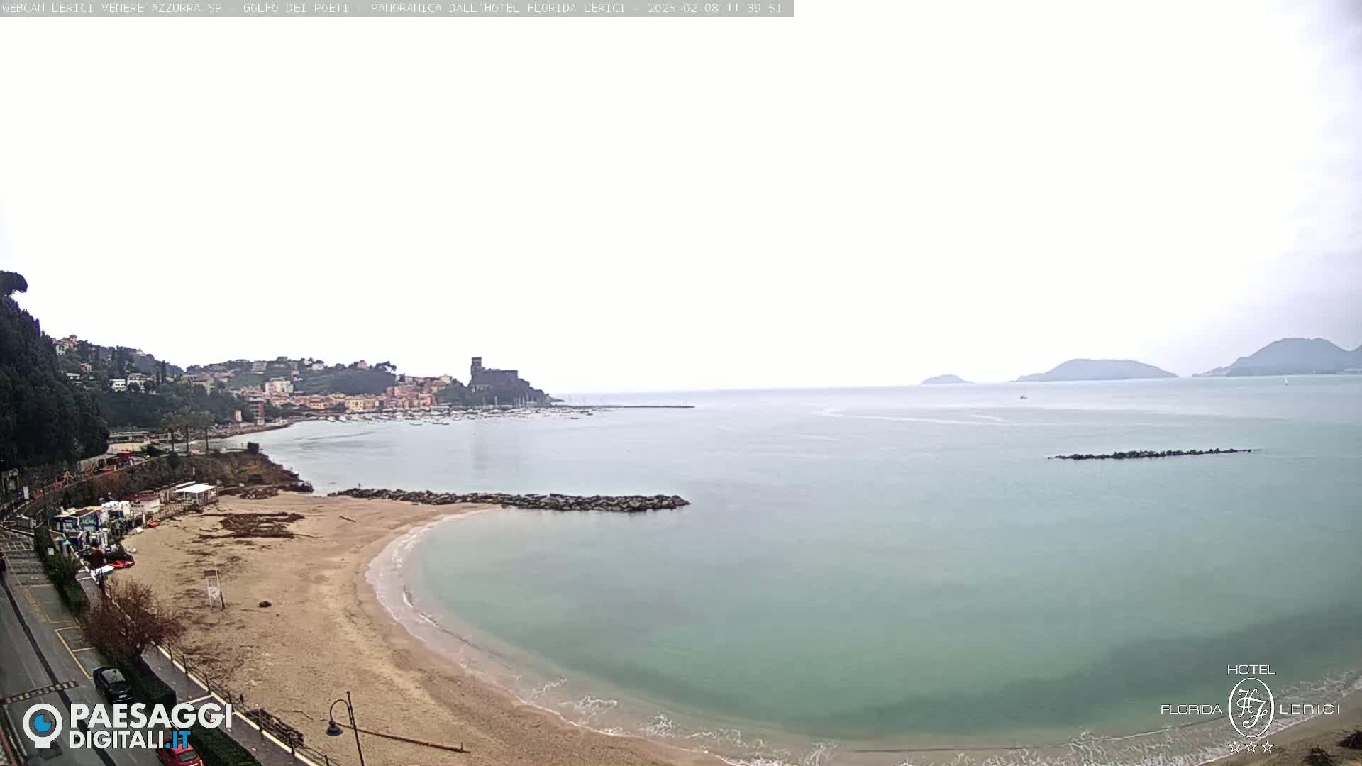 A sandy beach curves into a calm, light-green sea under an overcast sky, with a town and distant hills visible in the background.