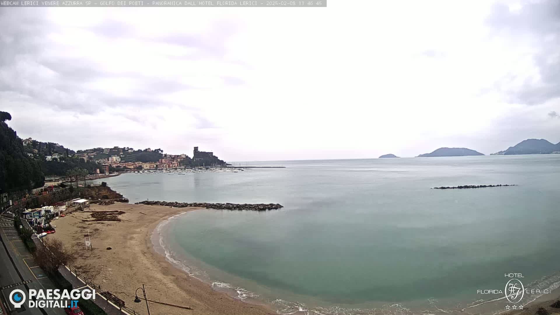 A sandy beach curves into a calm, blue-green sea under an overcast sky, with a town and distant hills visible across the water.