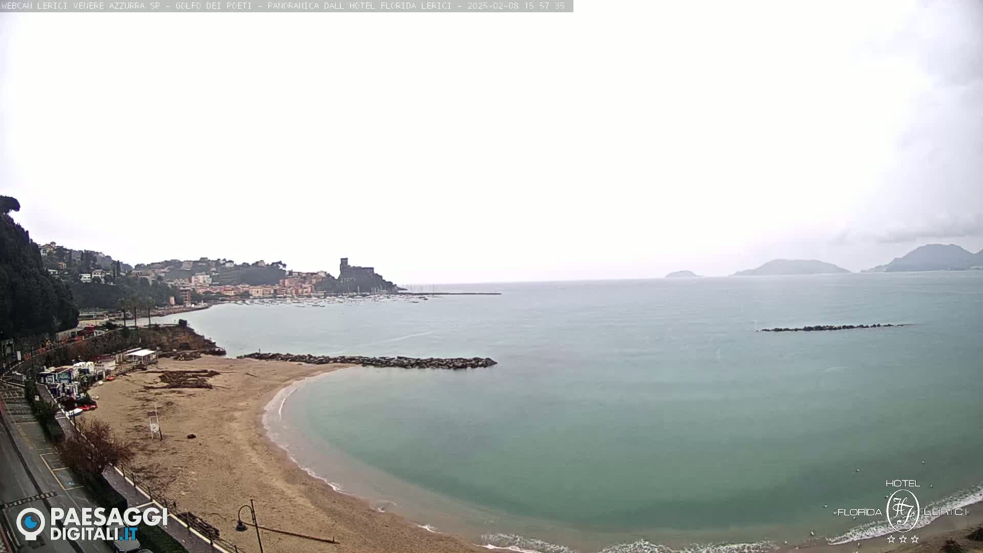 A sandy beach curves into a calm, teal-colored sea under an overcast sky, with a town and distant hills visible in the background.