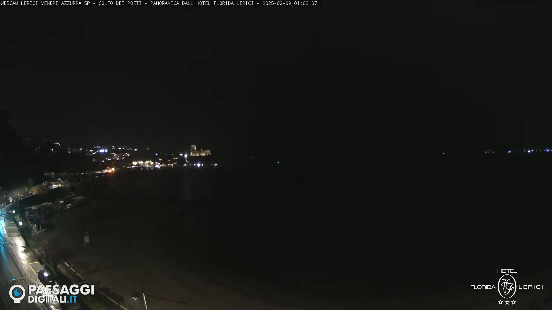 A nighttime, long shot of a dimly lit coastal town with a dark sea in the foreground.