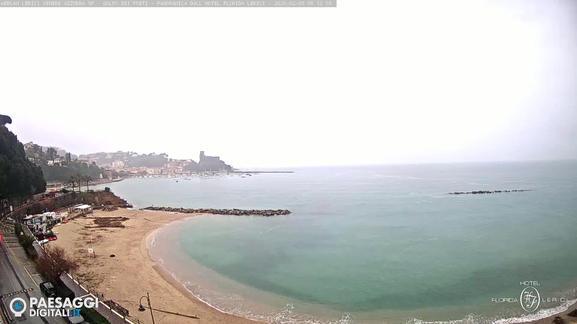 A hazy, overcast day reveals a sandy beach curving into a calm, teal sea, backed by a town nestled against a hillside.