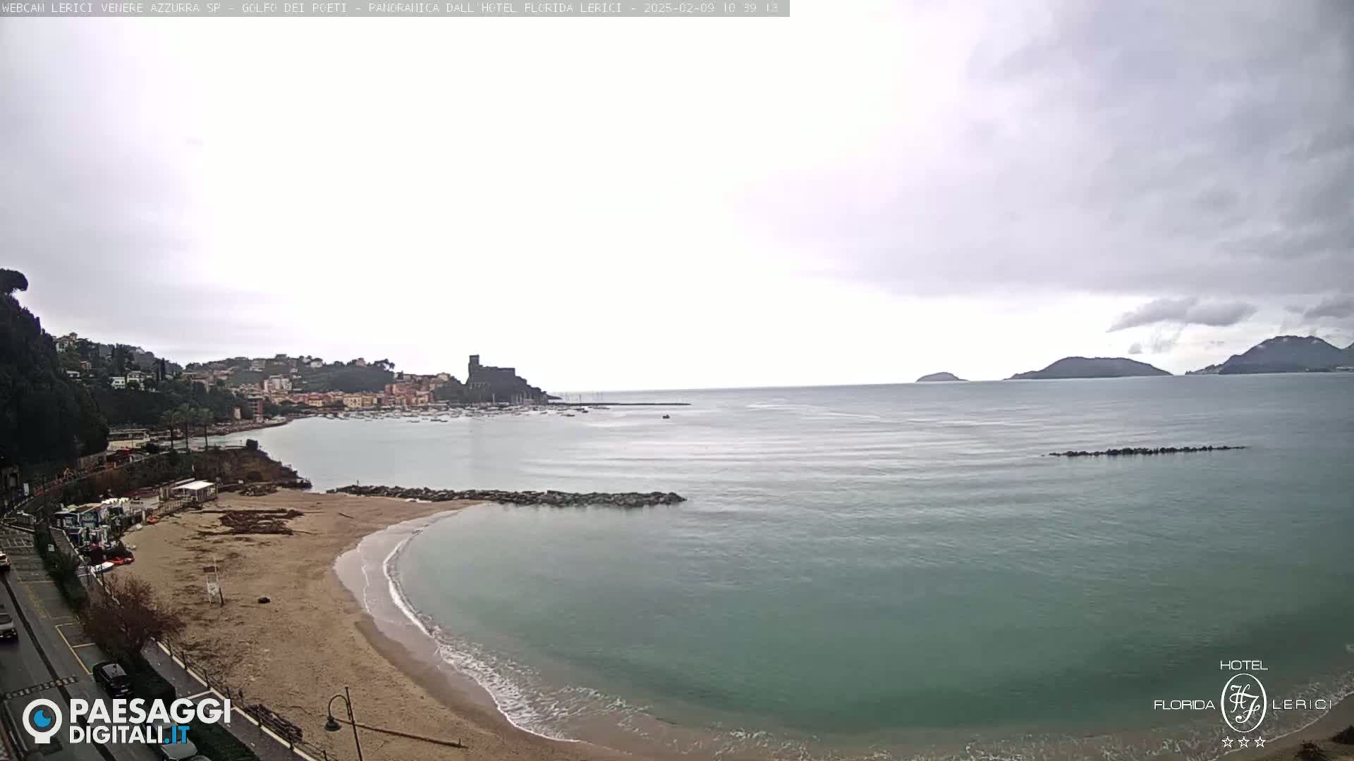 A beach with calm, light-green water under an overcast sky overlooks a town and distant hills.
