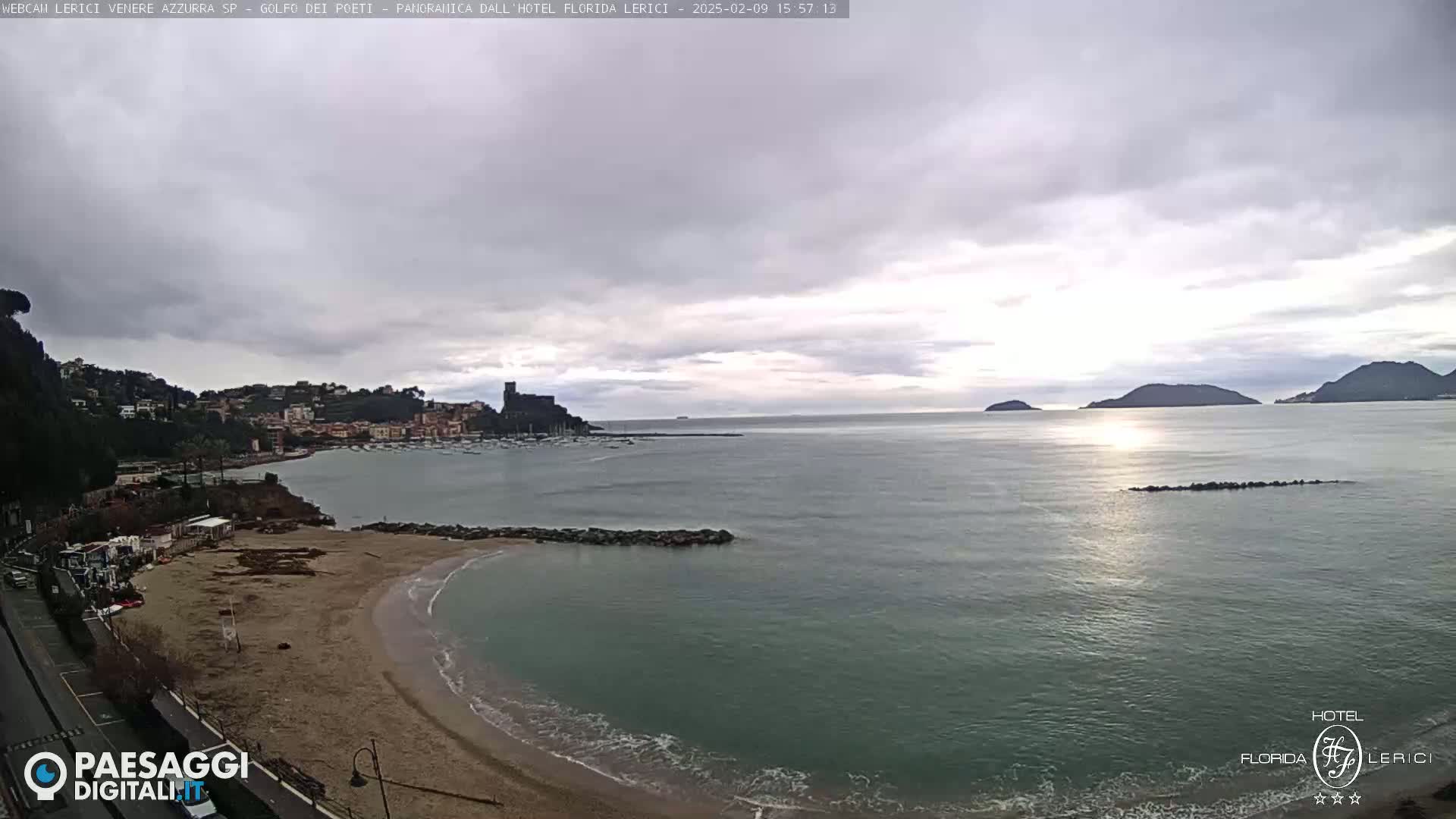 A sandy beach curves into a calm bay under a mostly cloudy sky, with a town and distant hills visible across the water.