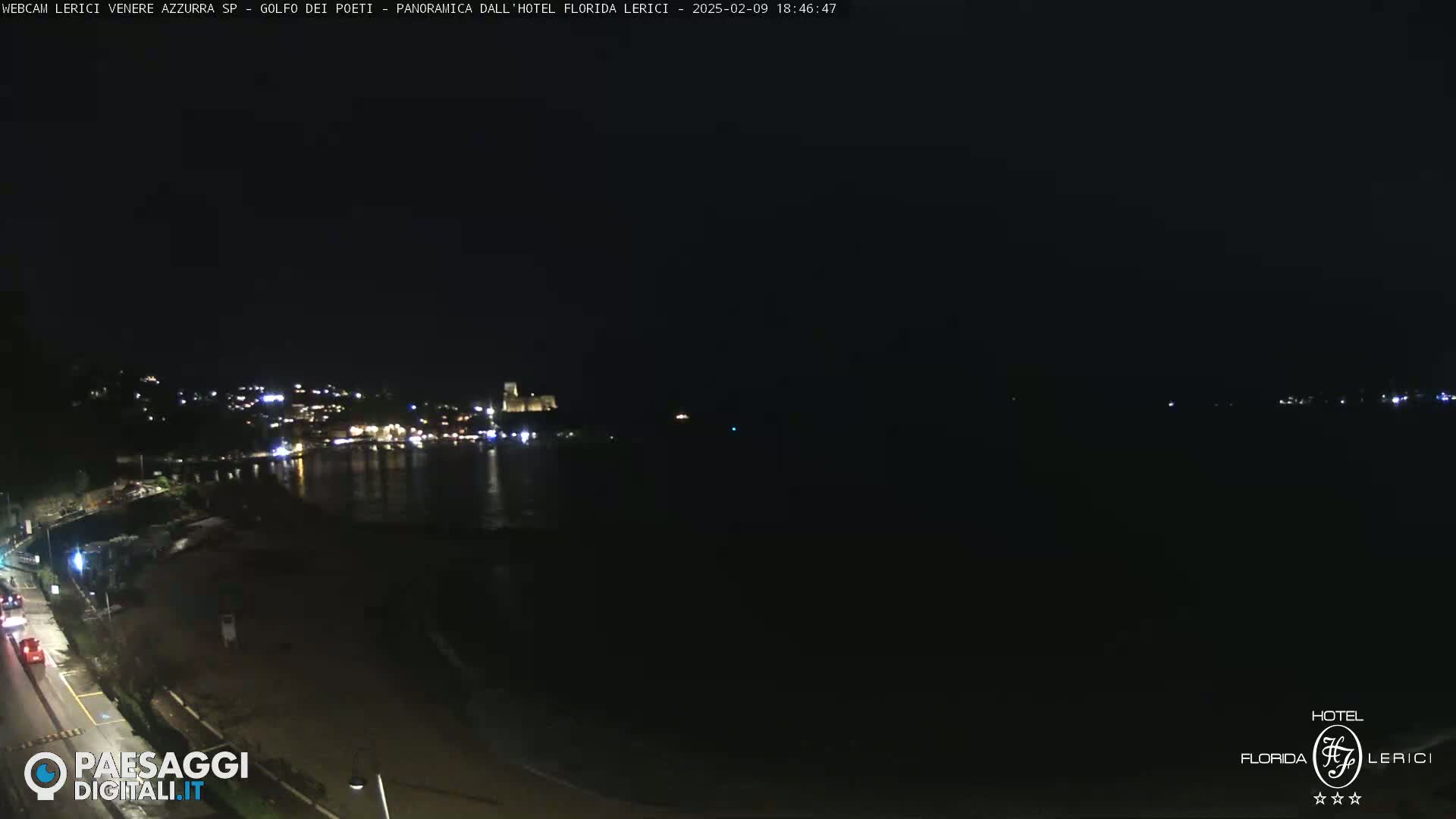 A nighttime coastal scene shows a dark beach and calm water reflecting dim lights from a town across the bay under a clear, dark sky.