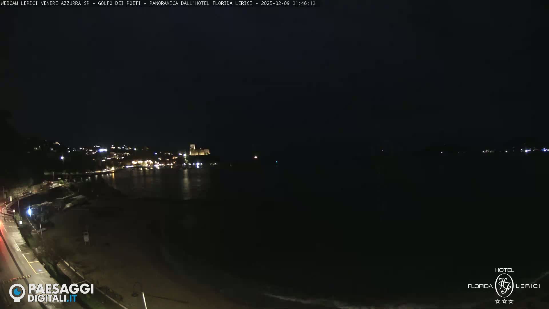 A nighttime view of a dark coastline with a town's lights visible across a calm body of water.