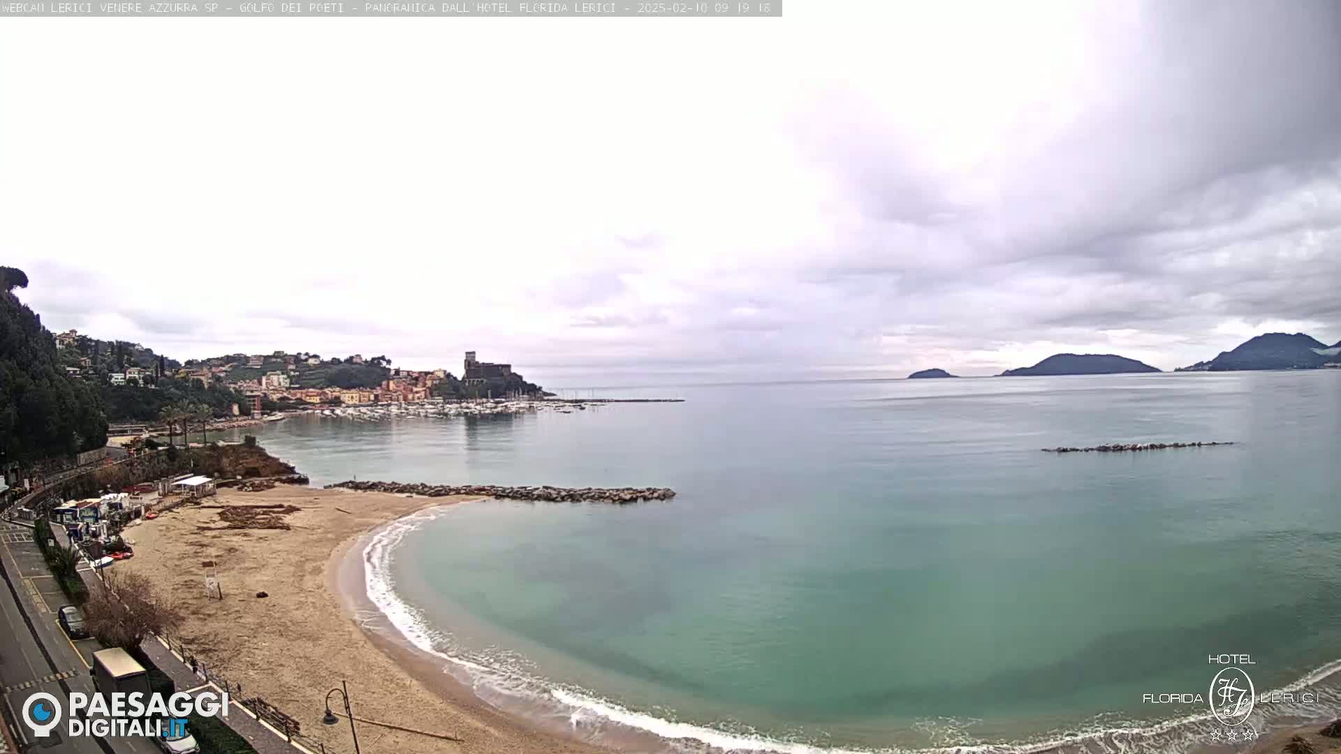 A sandy beach curves into a calm, teal sea under a cloudy sky, with a town and harbor nestled against a hillside in the background.
