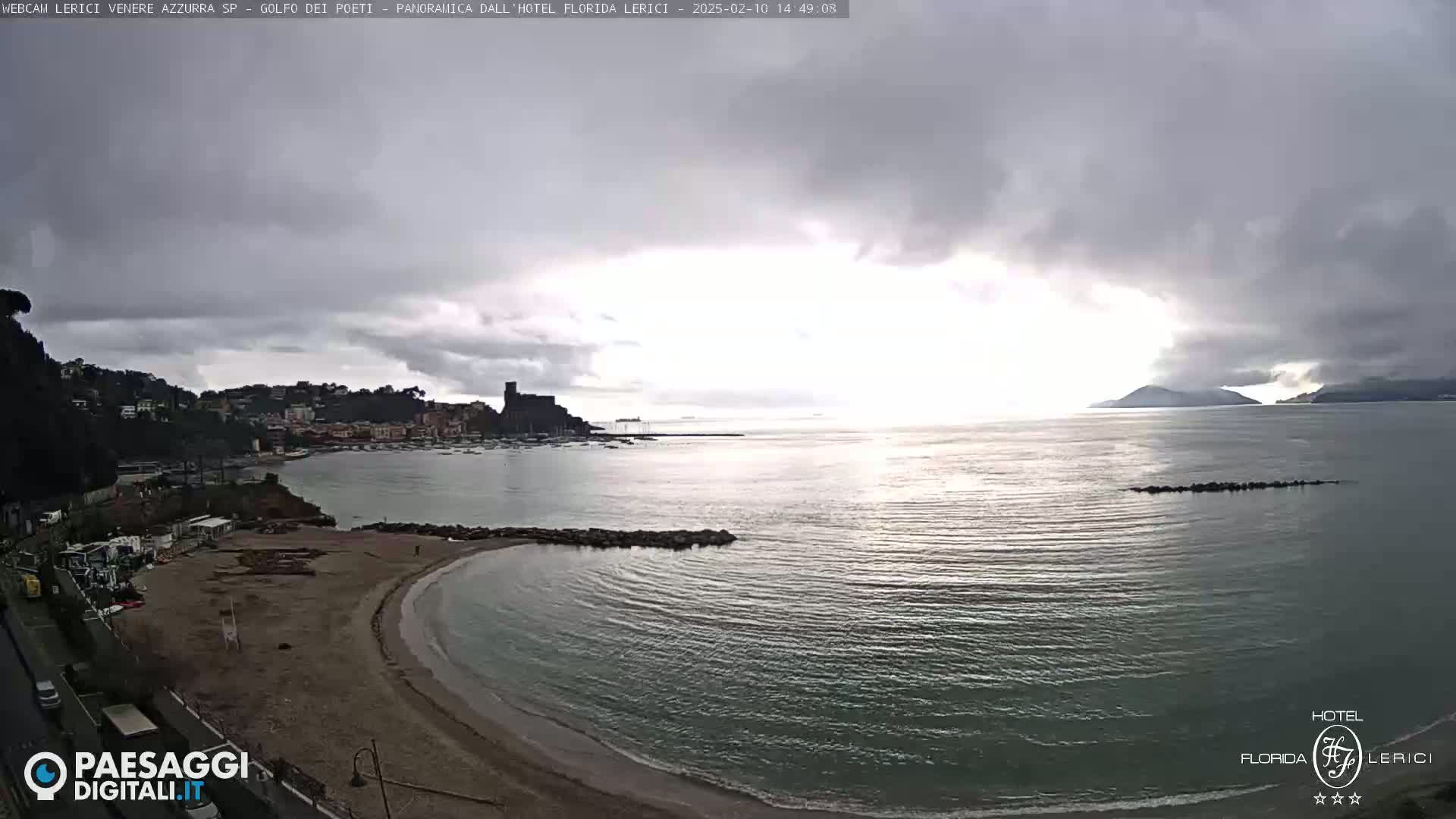 A mostly cloudy day reveals a sandy beach curving into calm, dark-green water, overlooking a town and distant islands.