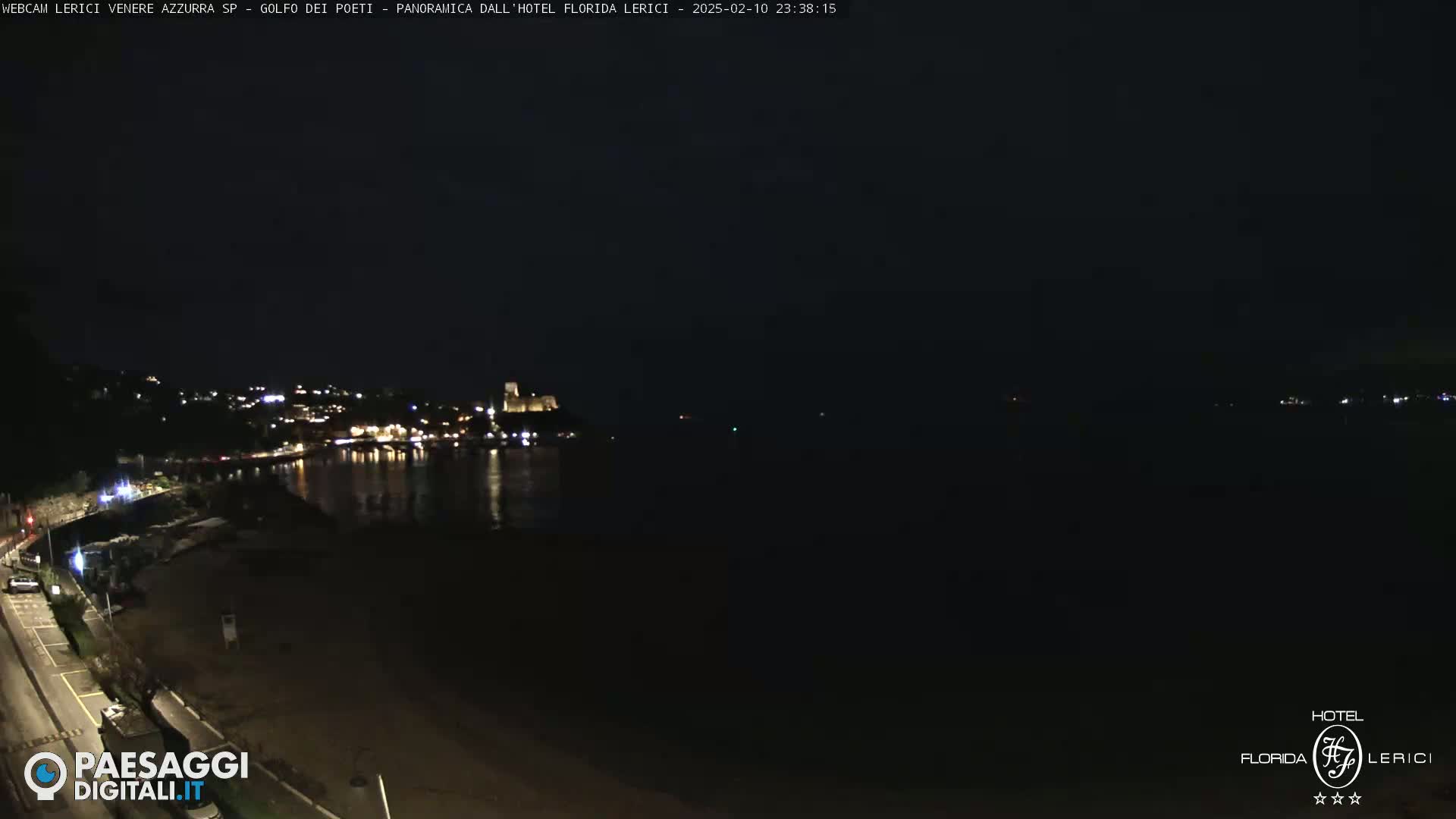 A nighttime coastal scene shows a dark beach and calm water reflecting the lights of a town across the bay, under a clear, dark sky.