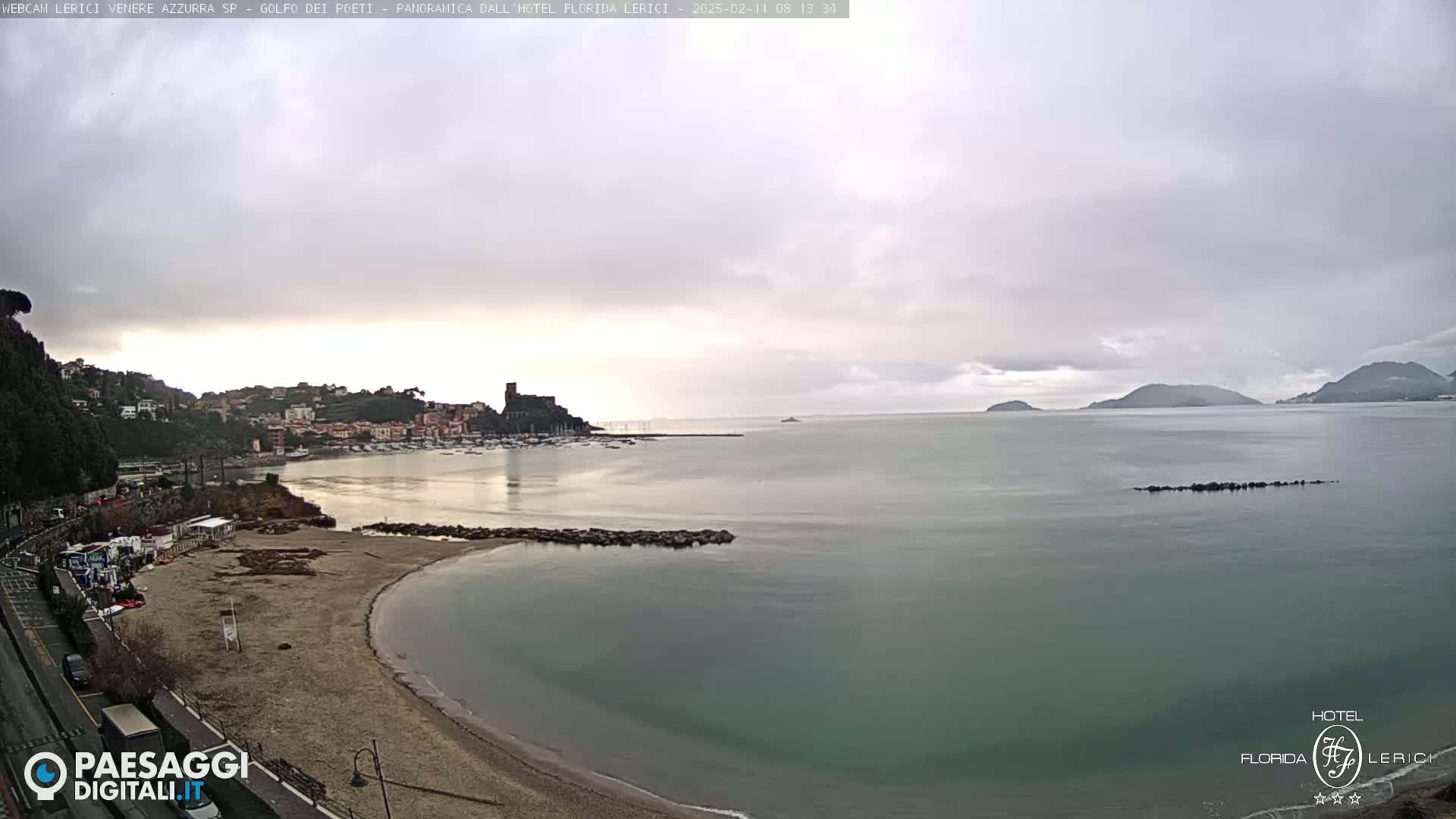 A beach with calm, greenish-grey water curves into a bay, under an overcast sky; hills and buildings are visible in the background.