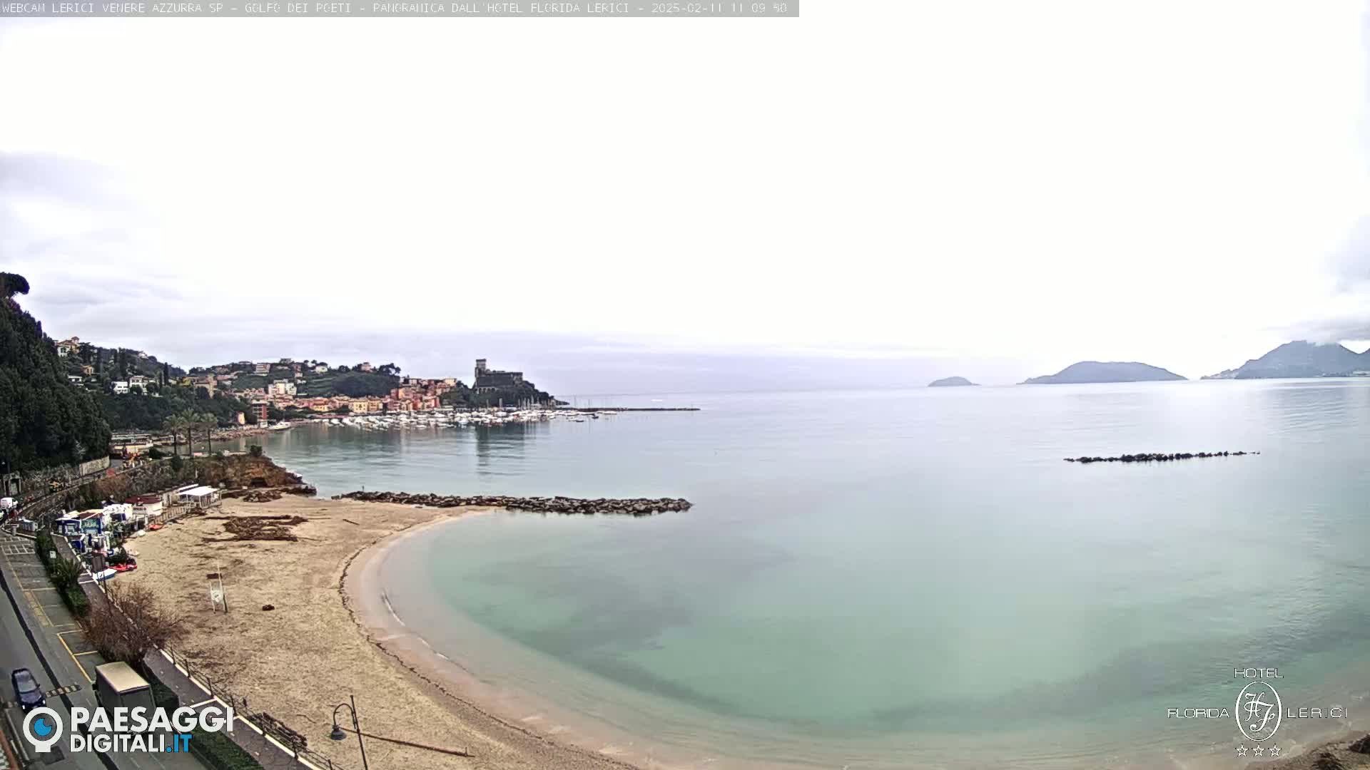 A sandy beach curves into a calm, pale-green sea under an overcast sky, with a town and harbor visible in the distance.
