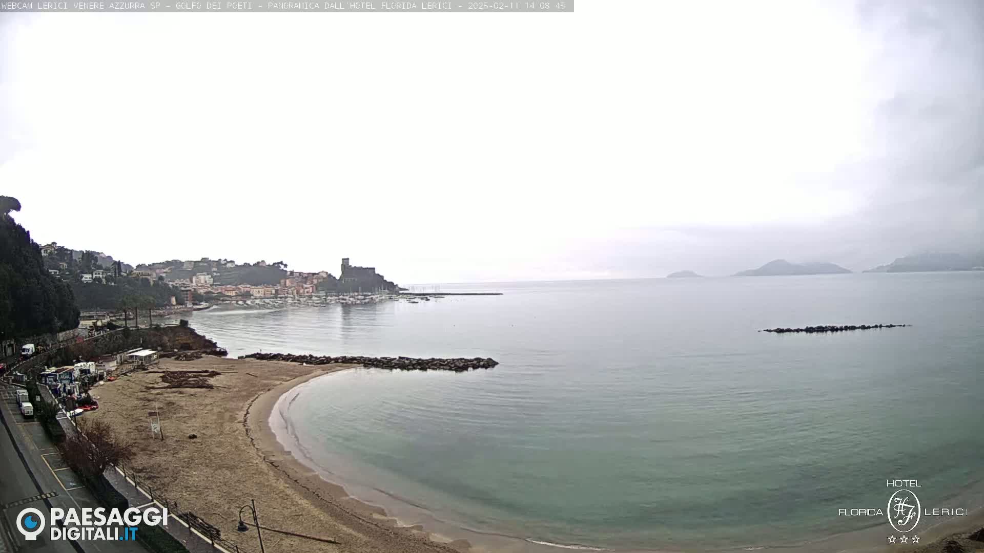 A mostly cloudy day shows a sandy beach curving into a calm sea, with a town and hills visible in the distance.