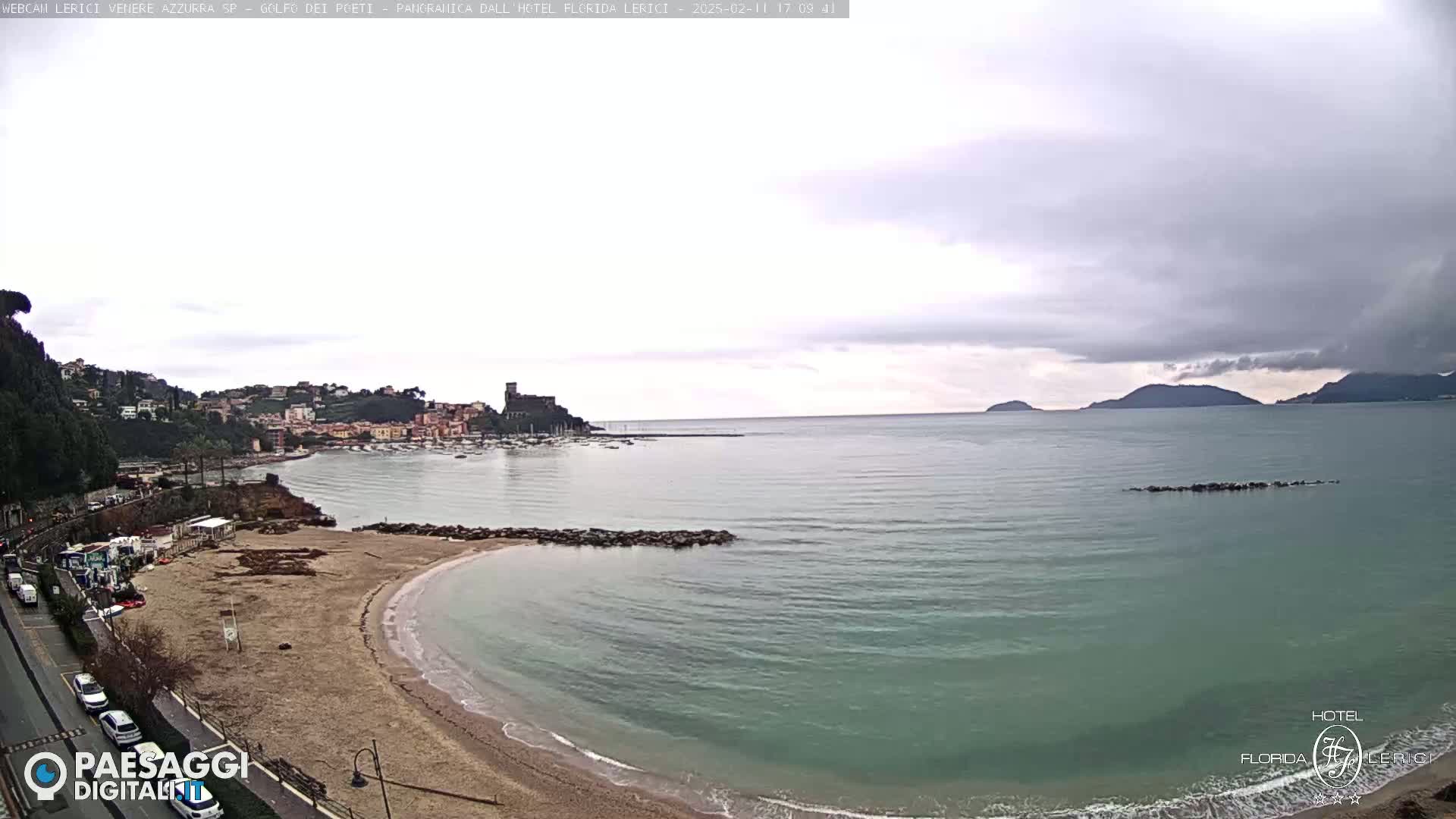 A coastal town with a sandy beach and calm, teal water is seen under an overcast sky.
