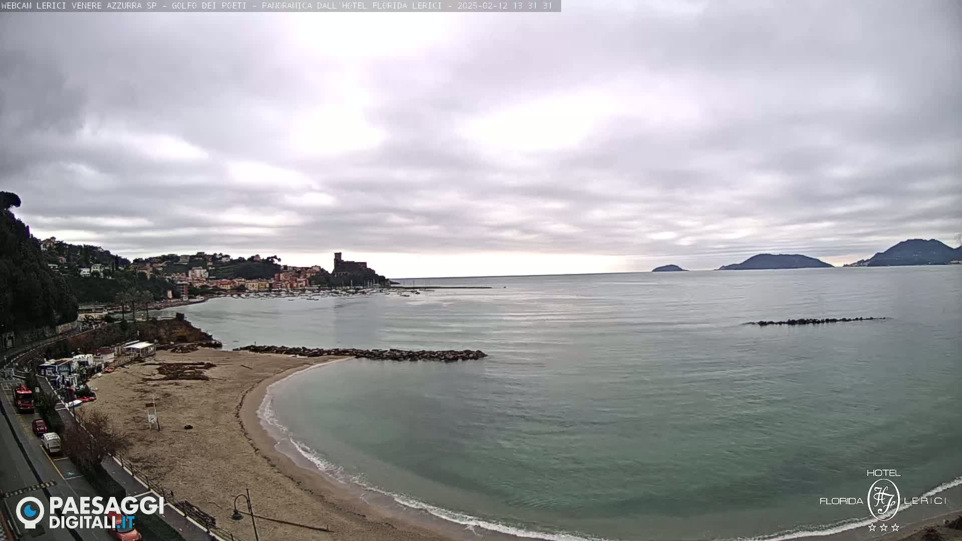 A sandy beach curves into a calm, greenish-grey sea under a cloudy sky, with a town and distant hills visible in the background.