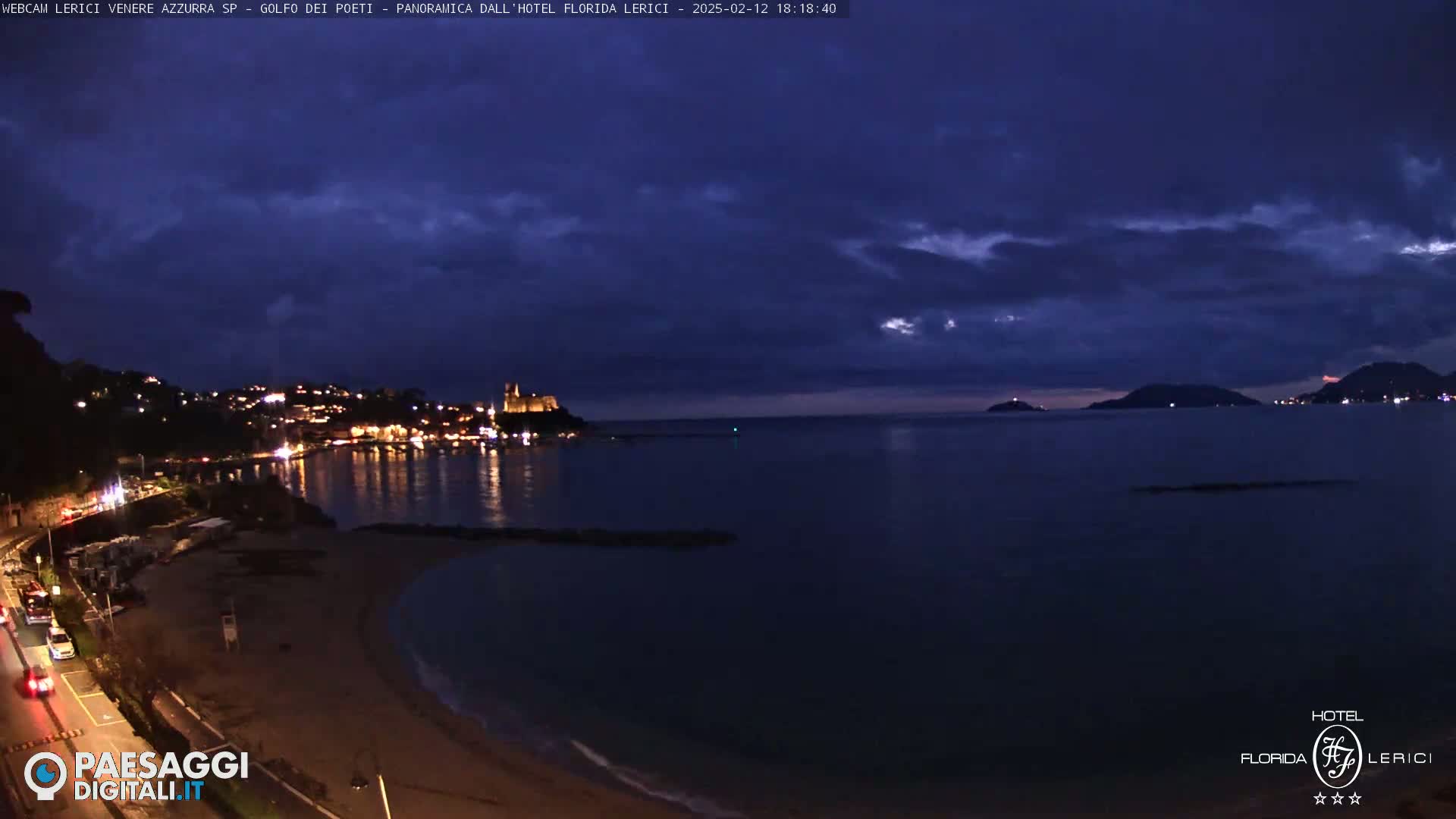 A nighttime coastal scene shows a dark, cloudy sky over a calm sea with a town's lights illuminating the shoreline and a castle in the distance.
