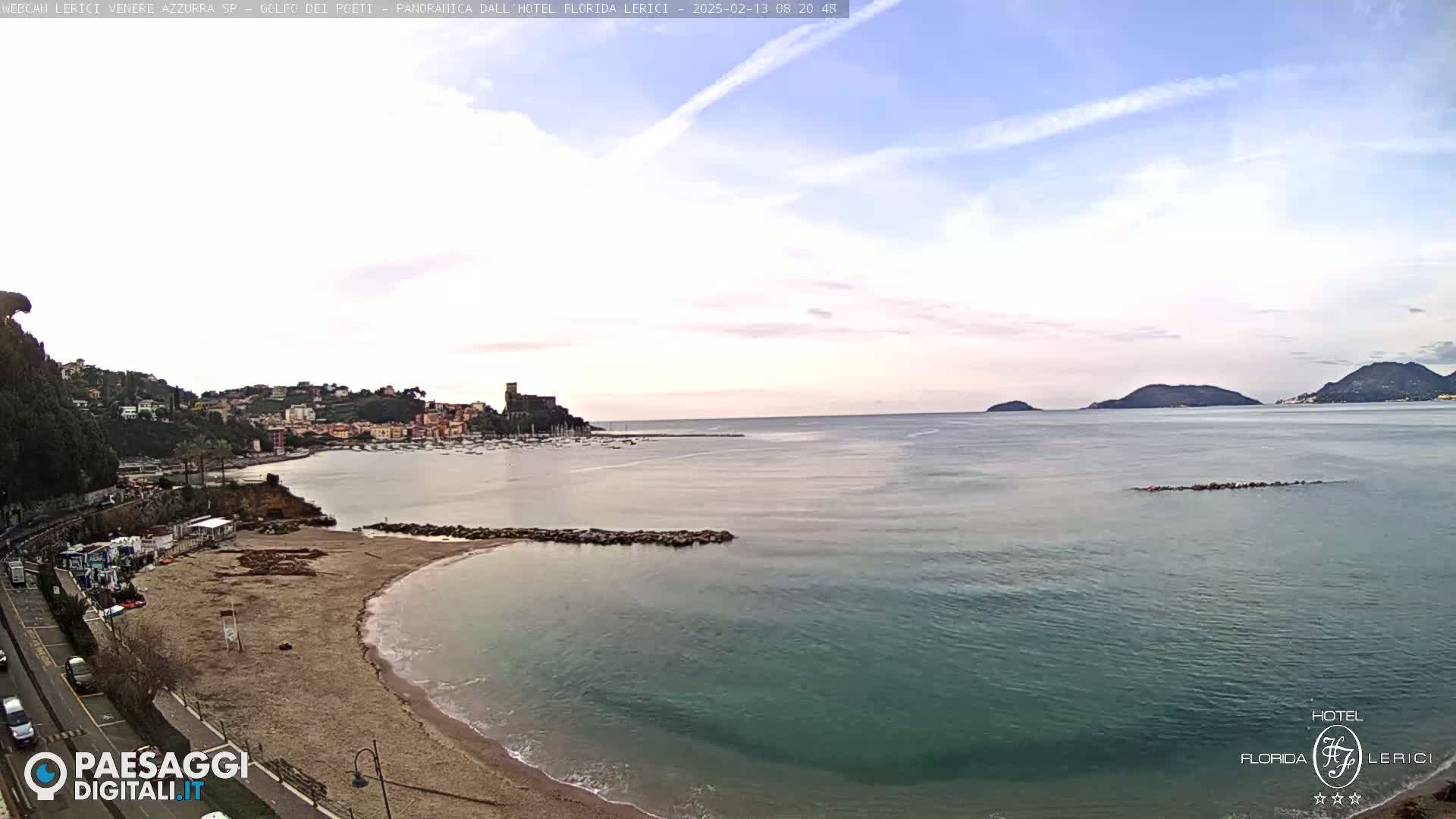 A sandy beach curves into a calm, teal sea, with a town and distant islands visible under a mostly clear sky.