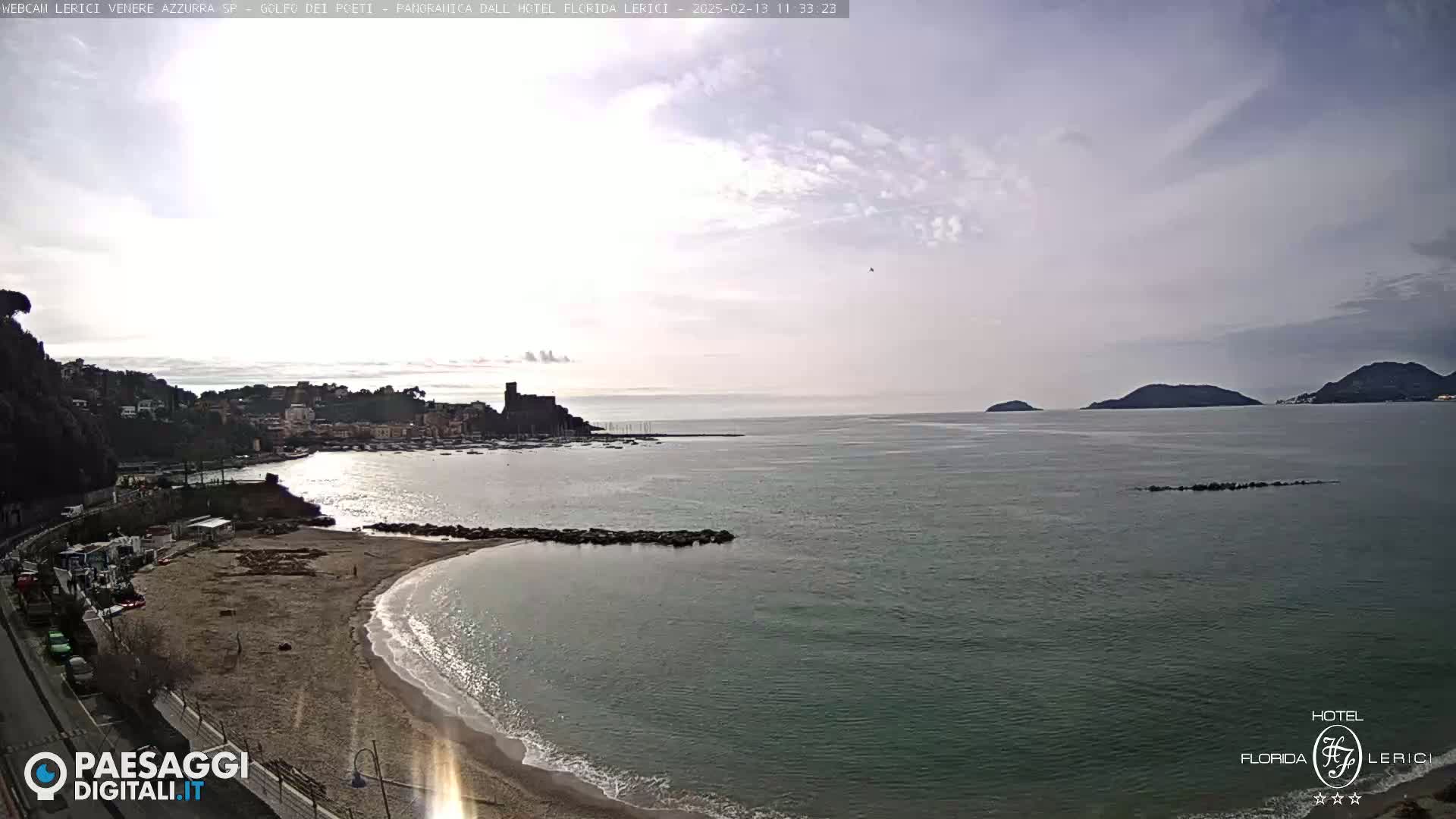 A sandy beach curves into a calm bay, overlooking a town and distant islands under a partly cloudy sky.