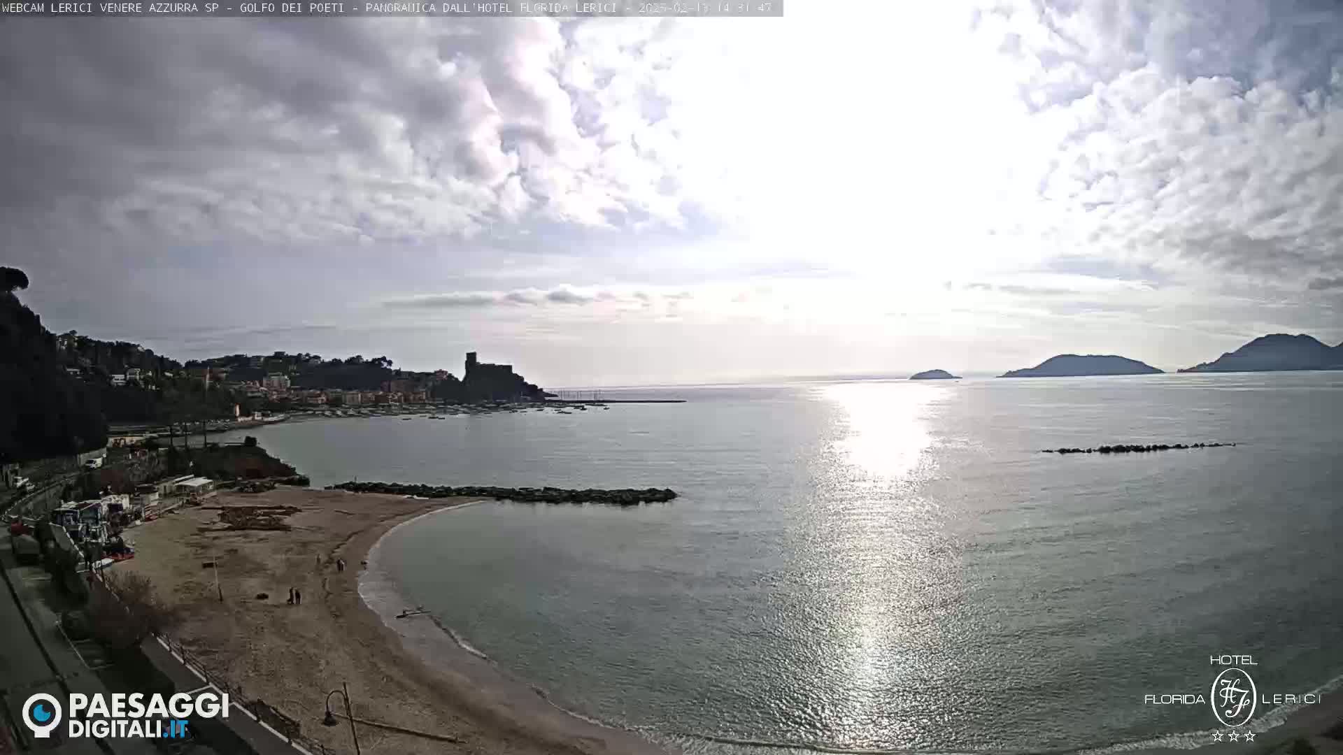 A partly cloudy day shows a sandy beach curving into a calm bay, with a town and distant hills visible across the water.