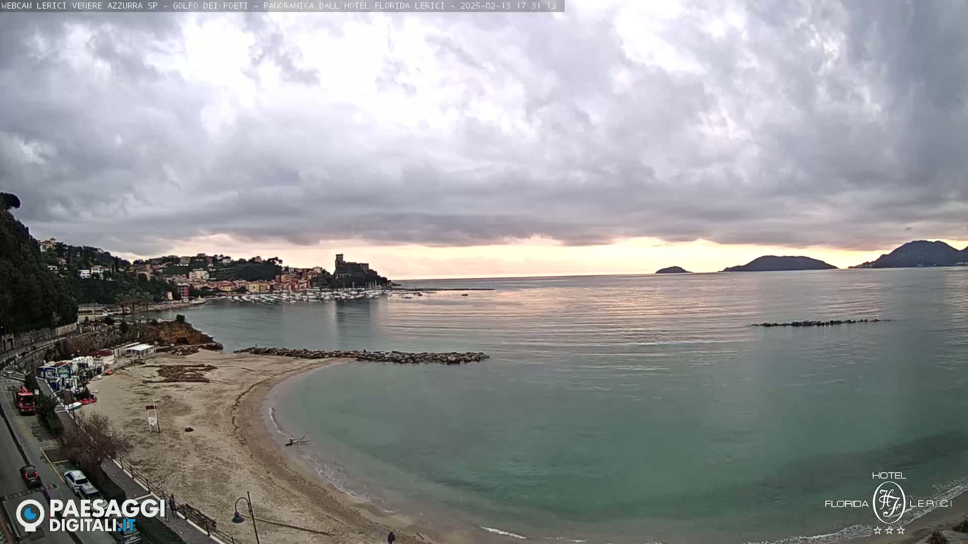 A mostly cloudy sunset over a calm bay with a sandy beach and a town nestled on a hillside.