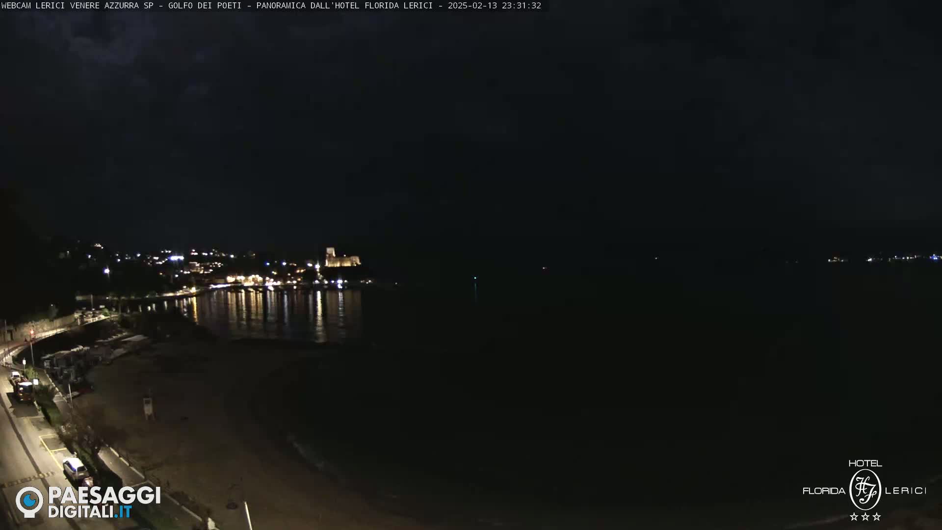 A nighttime coastal town scene with a dark sky and calm water reflecting the lights of buildings along the shore.