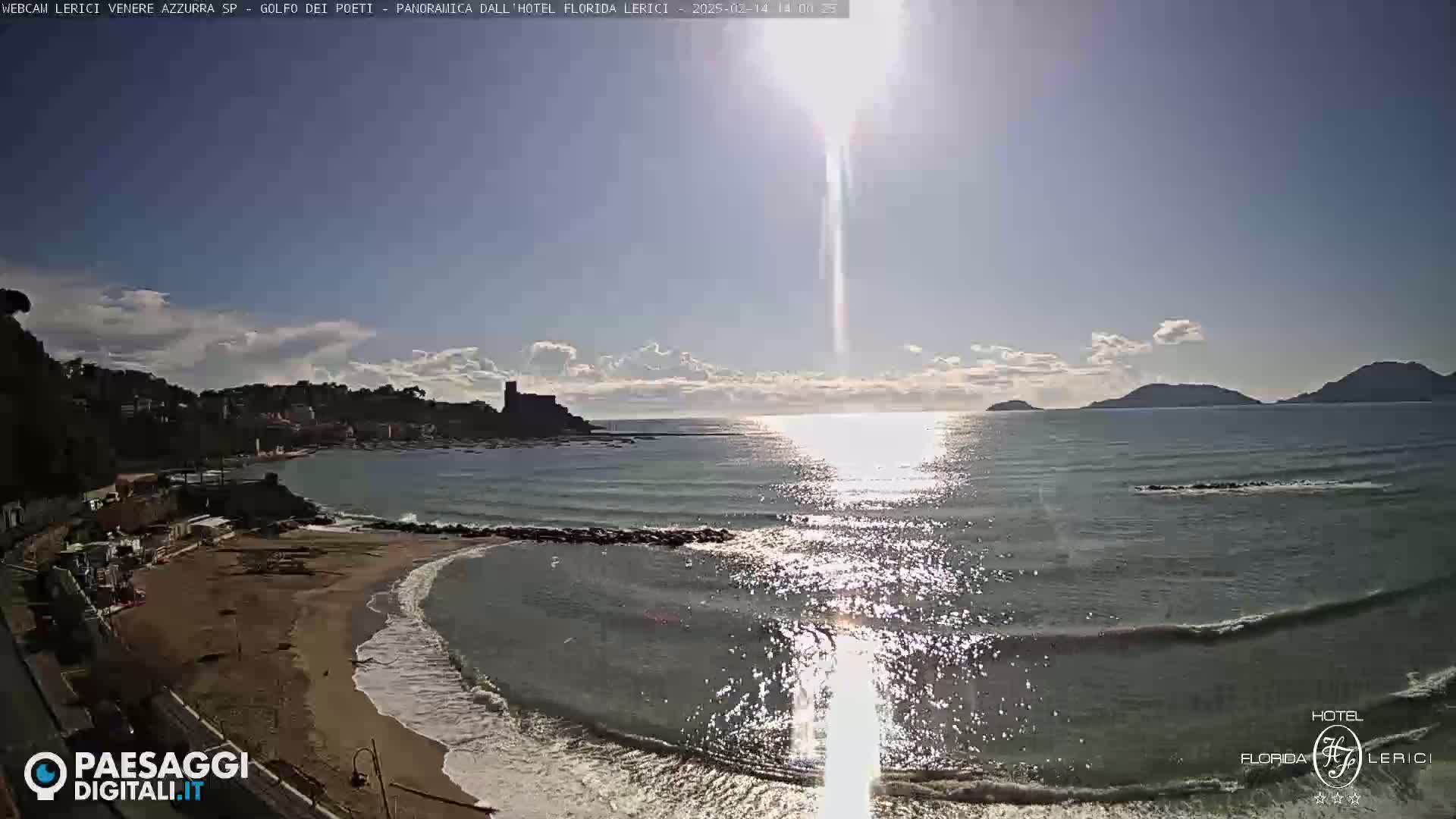 A sandy beach curves into a calm sea under a mostly sunny sky, with a distant castle and hills visible in the background.