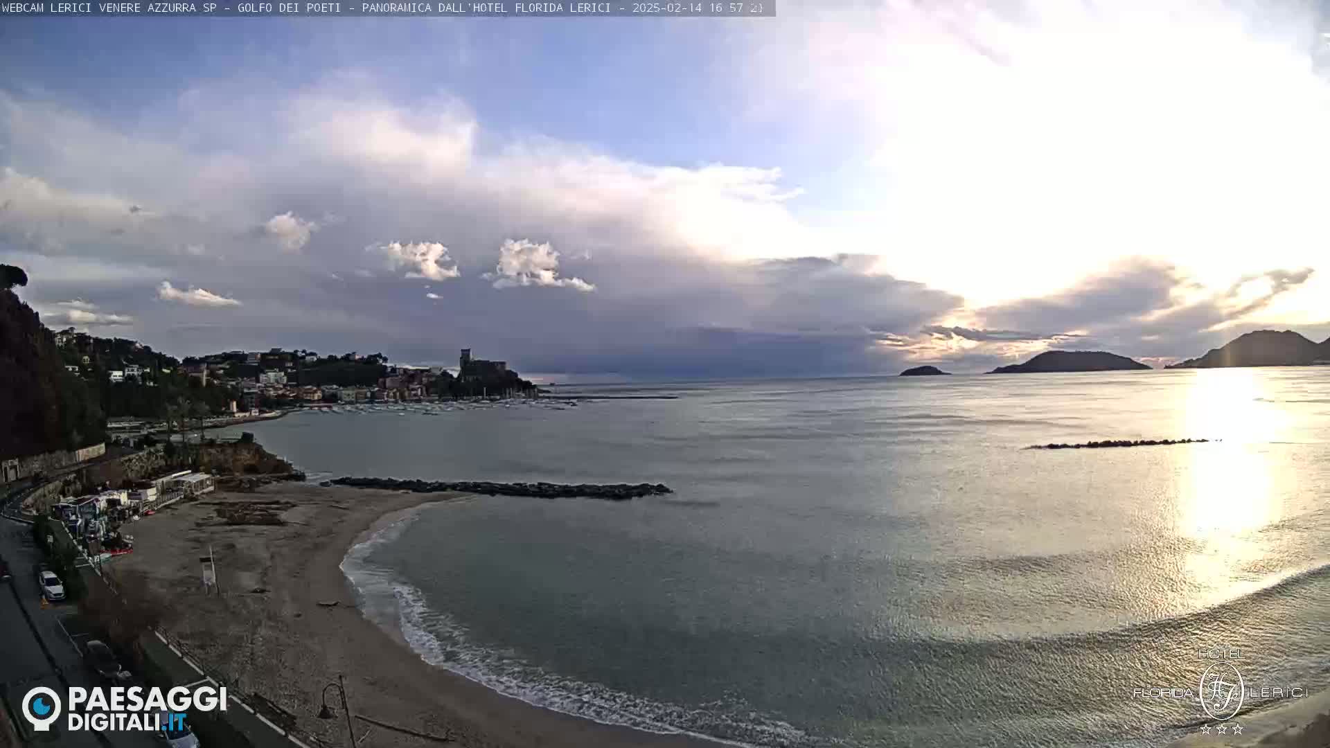 A partly cloudy sunset over a calm bay with a sandy beach, a town, and distant hills.