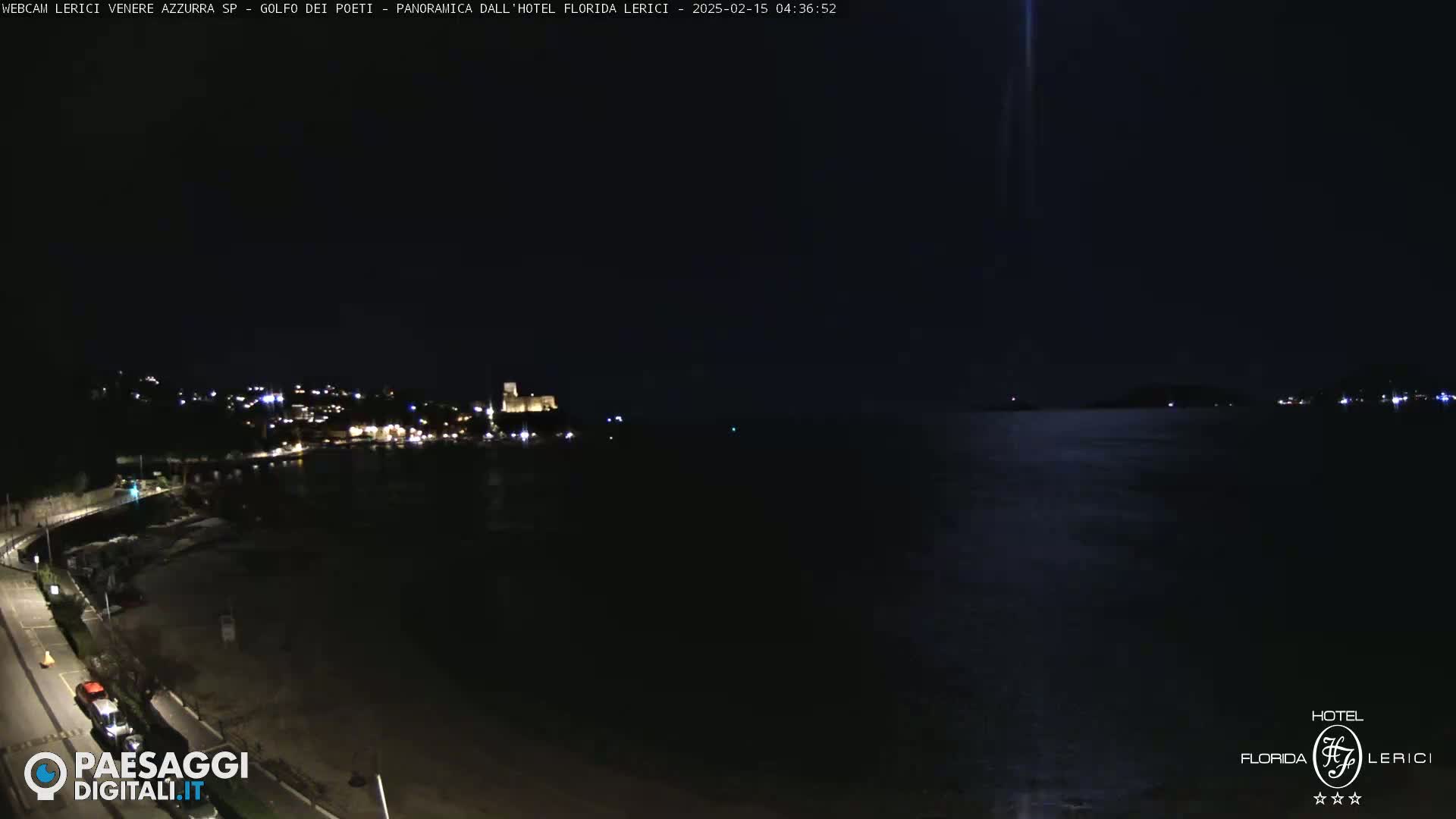 A nighttime coastal scene shows a dark body of water with a dimly lit town and a castle on the opposite shore, under a clear, dark sky.