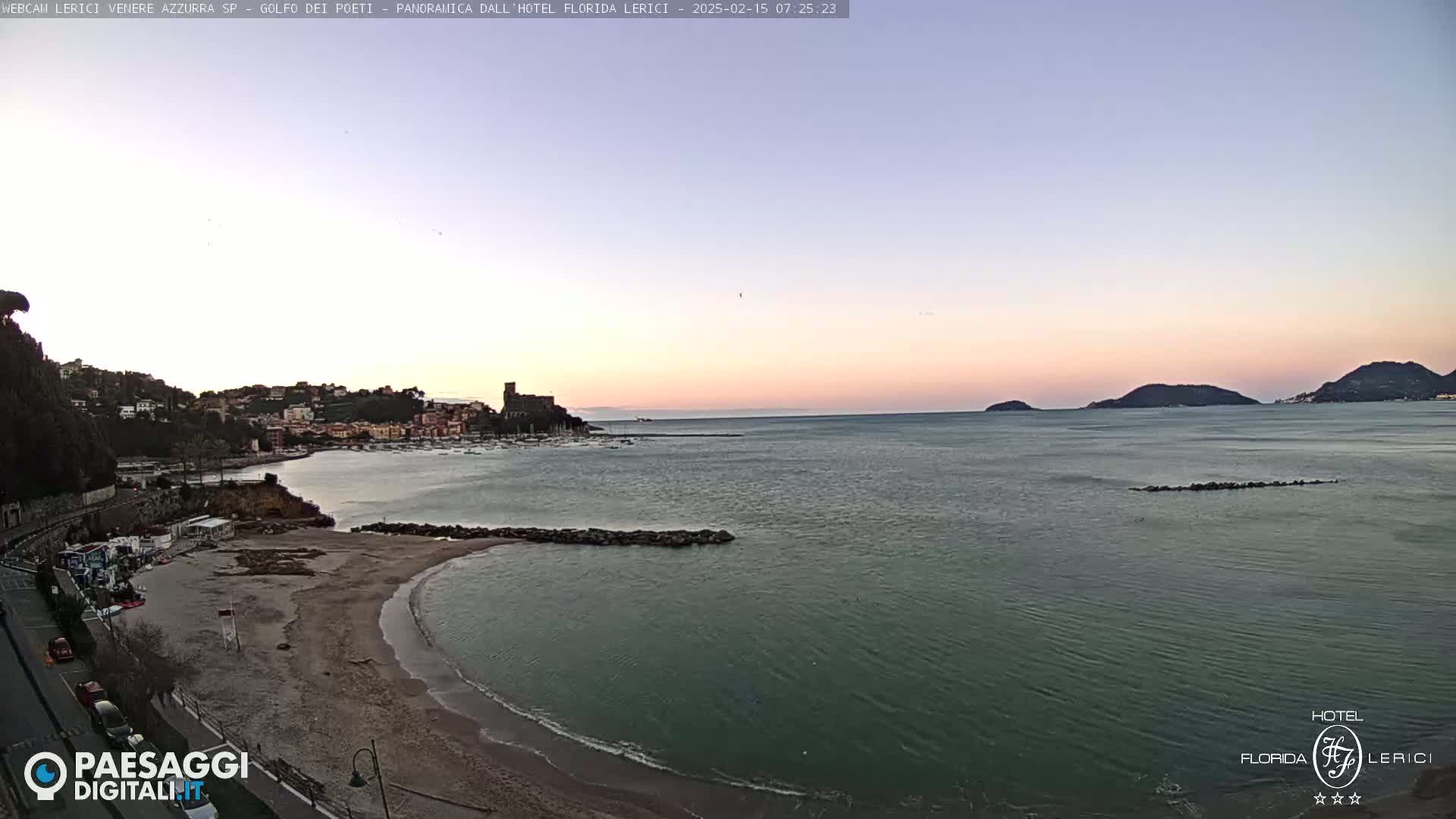 A tranquil coastal scene at dawn shows a sandy beach curving into a calm sea, with a town nestled on a hillside in the background and islands visible in the distance under a pale pink and blue sky.