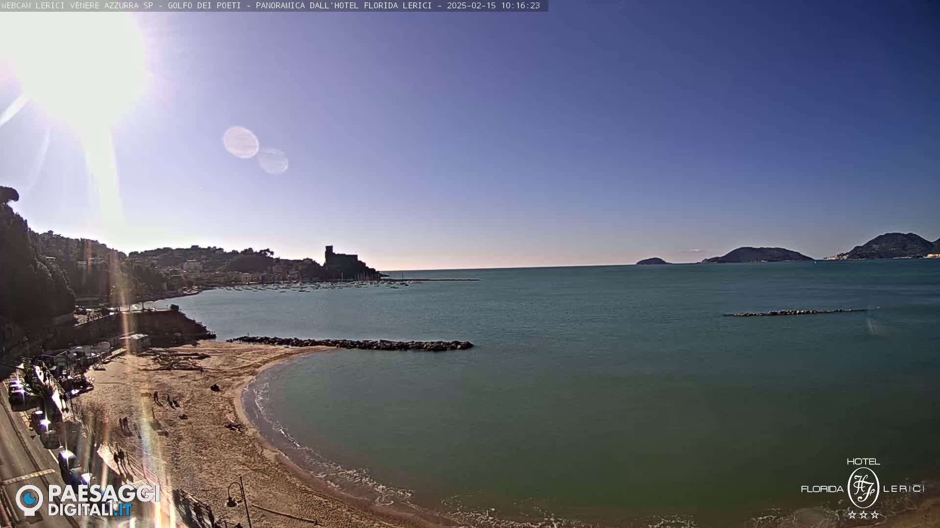 A sandy beach curves into a calm, teal sea under a clear blue sky, with a town and distant hills visible across the water.