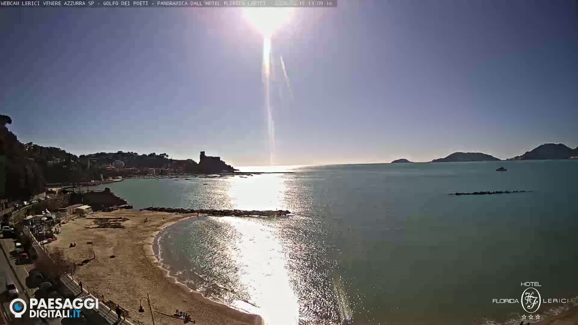 A sunny day reveals a sandy beach, calm sea, and a town nestled amongst hills, with a distant island visible on the horizon.