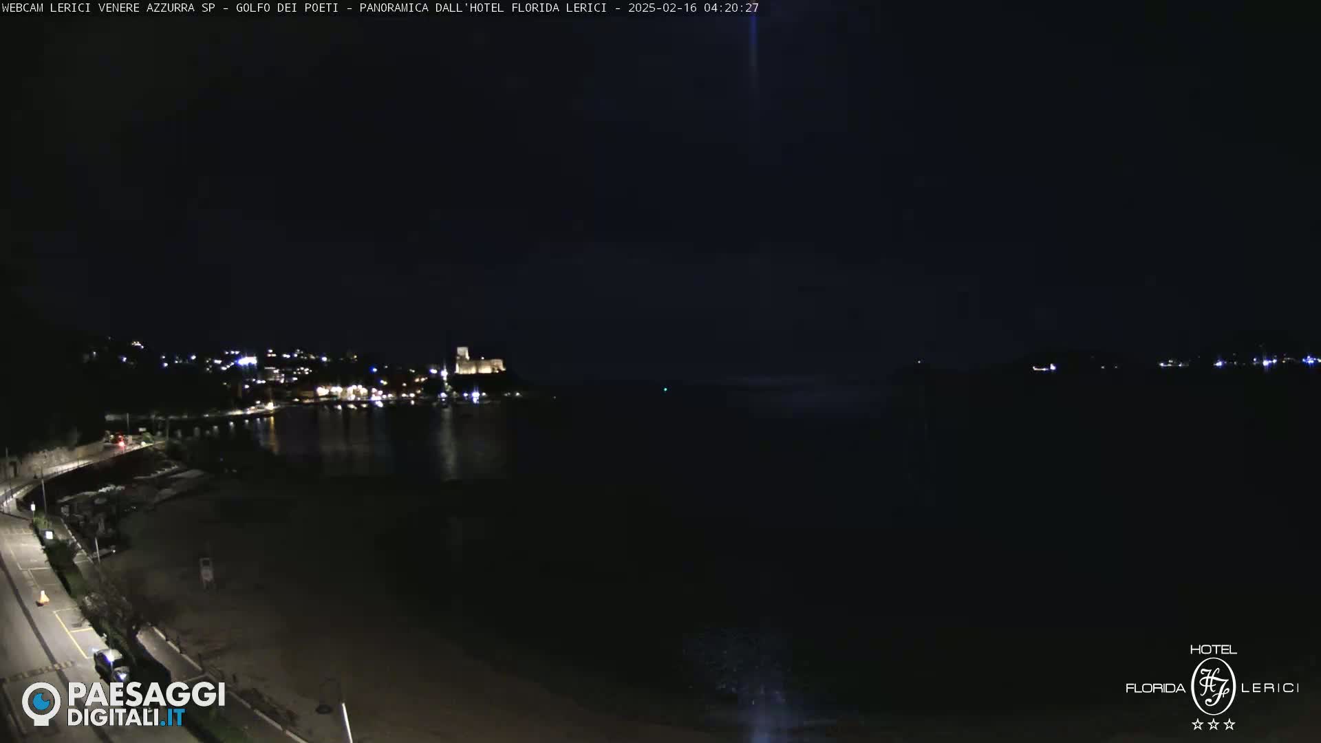 A nighttime coastal scene shows a dark beach, calm water, and a small town with lights on a hill across the water.