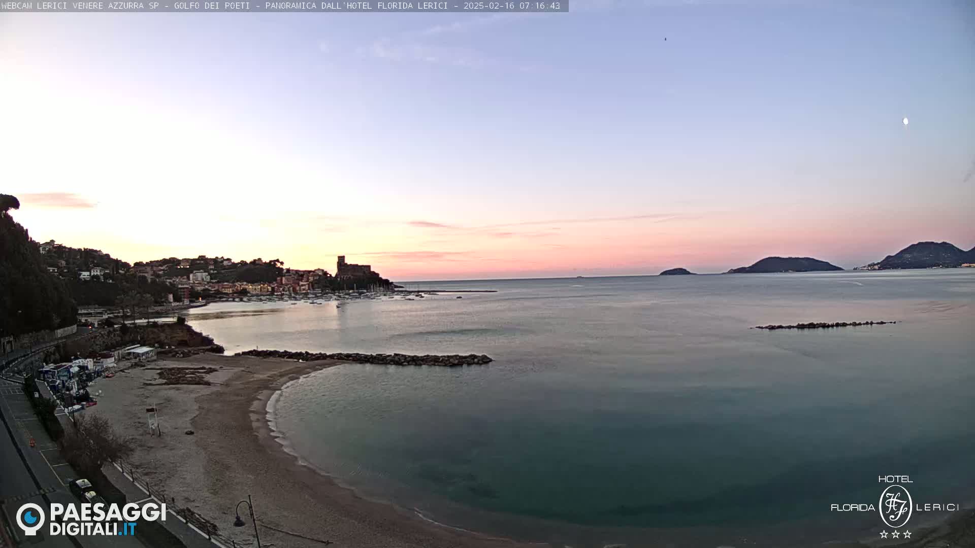 A calm, sandy beach curves into a bay at sunset, with a town nestled on a hillside across the water, under a pale pink sky.