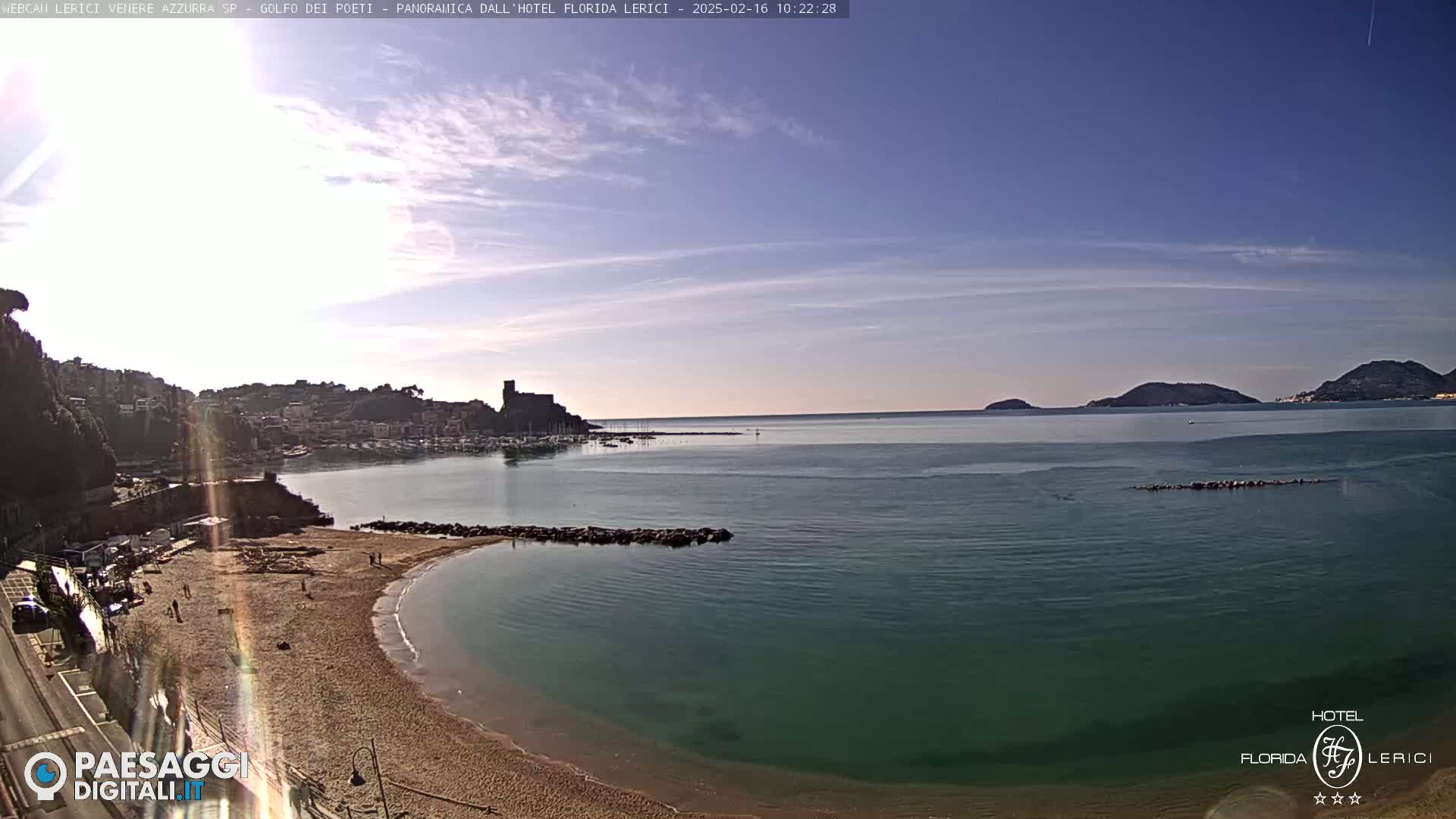 A sandy beach curves into a calm, teal-colored bay, overlooking a town and harbor with hills and islands in the background under a mostly clear sky.